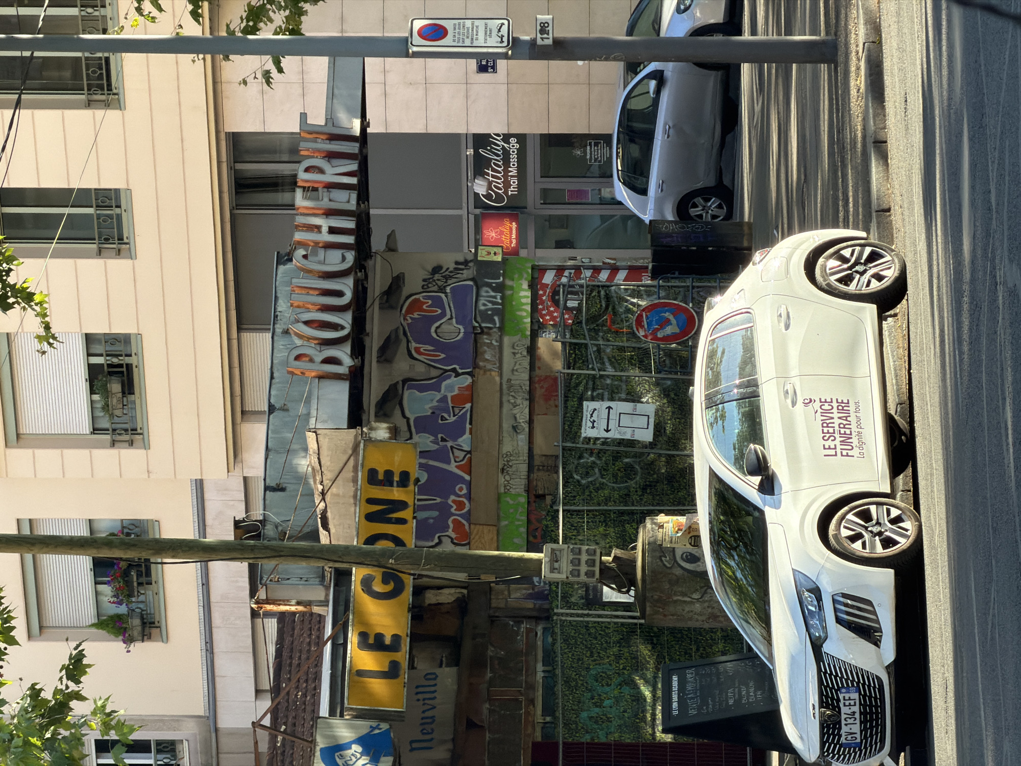 The image shows a street scene in Lyon, France. In the foreground, a white Peugeot 208 is parked on the street. The car has "LE SERVICE FUNÉRAIRE La dignité pour tous" written on its side. Behind the car, there are several shops, including one with a neon sign that reads "BOUCHERIE". Another sign reads "LE GONE". Further back, a silver car is parked. The street is lined with buildings and trees. The overall impression is of a typical urban street with shops and parked cars.