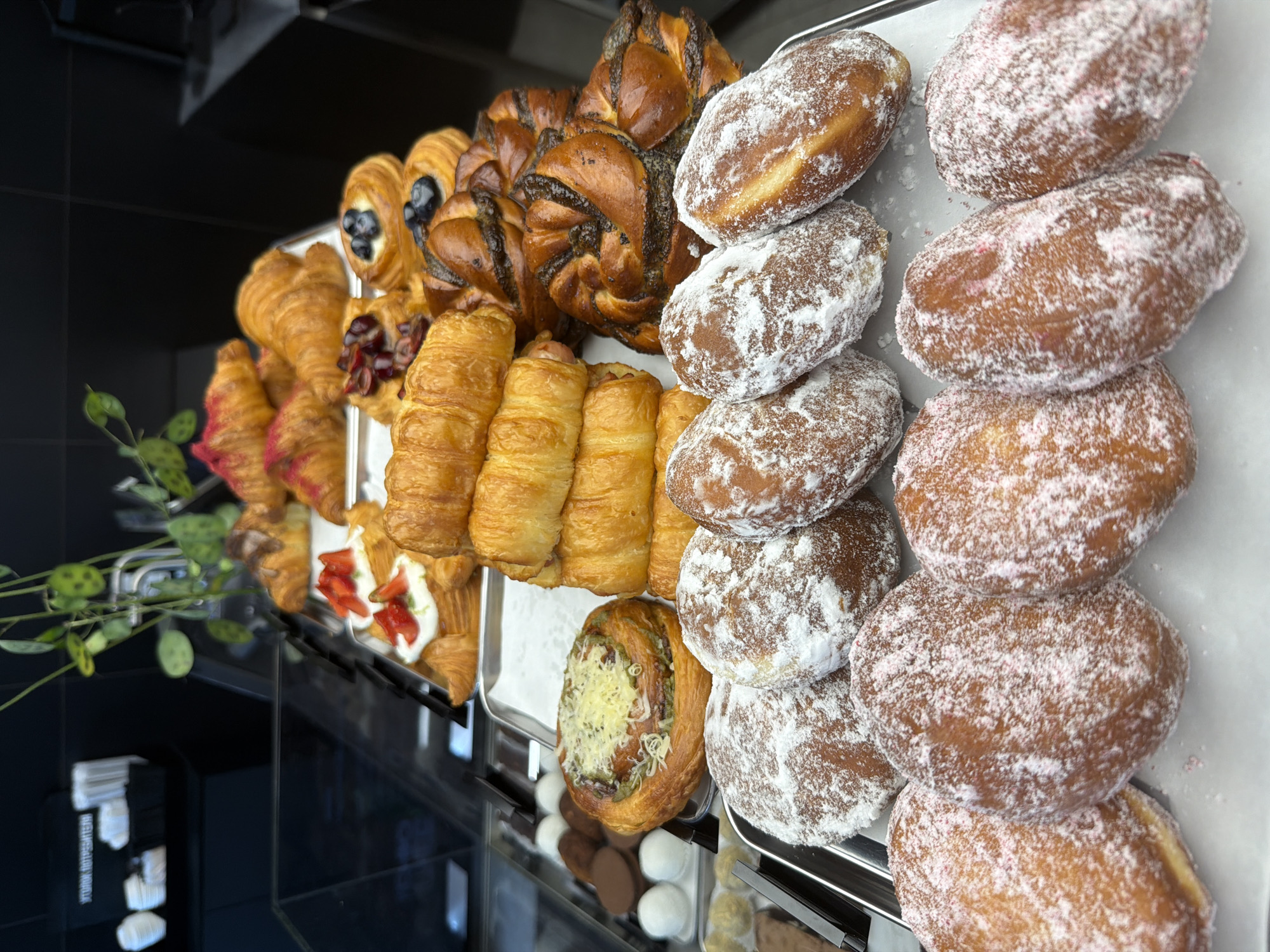 The image shows a display of assorted pastries in what appears to be a bakery or cafe. The pastries are arranged on metal trays and in a glass display case. Visible items include croissants, some topped with strawberries and cream, others with pink frosting. There are also rolled pastries, buns with poppy seeds, and round donuts dusted with powdered sugar, some with pink sprinkles. In the background, there are more pastries in a refrigerated case, along with some cups and what look like napkins on a shelf. A green plant is visible in the upper right portion of the image. The lighting suggests an indoor commercial setting. The location is identified as Izhevsk, Russia. There are no people visible in the image.