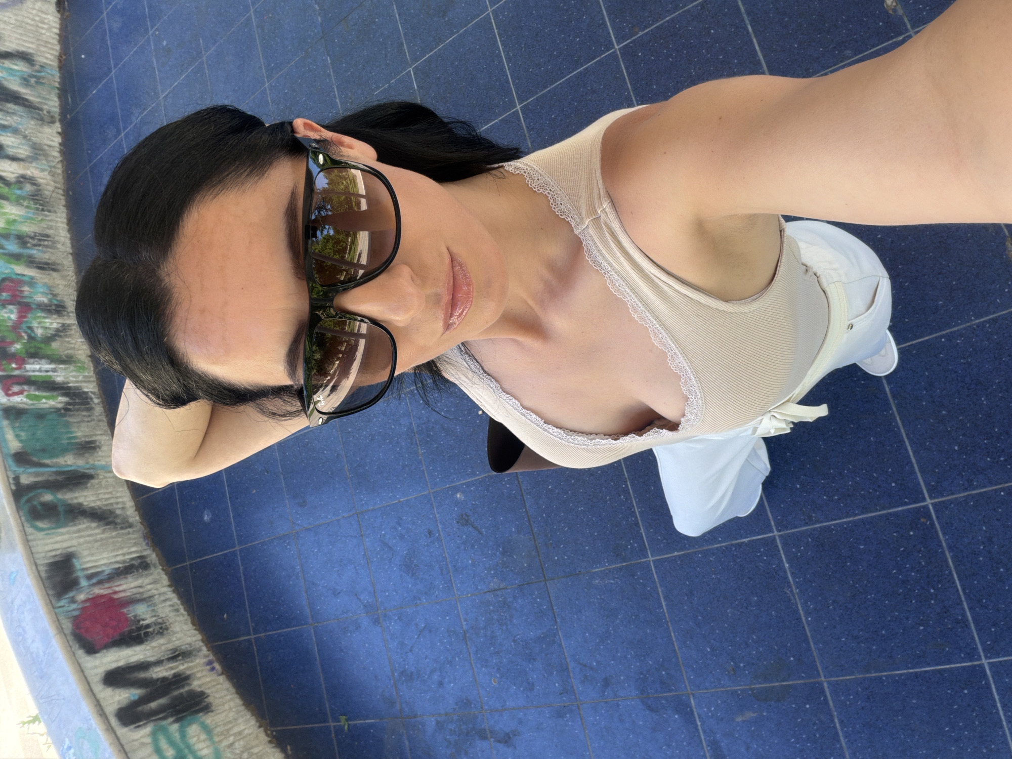A woman is taking a selfie outdoors. She has dark hair and is wearing sunglasses, a tan tank top with lace trim, and white pants. The background appears to be a public area with blue tiled flooring and a curved concrete wall with graffiti. The reflection in her sunglasses shows trees and a clear sky. The scene is set in the city of Pleven, Bulgaria.