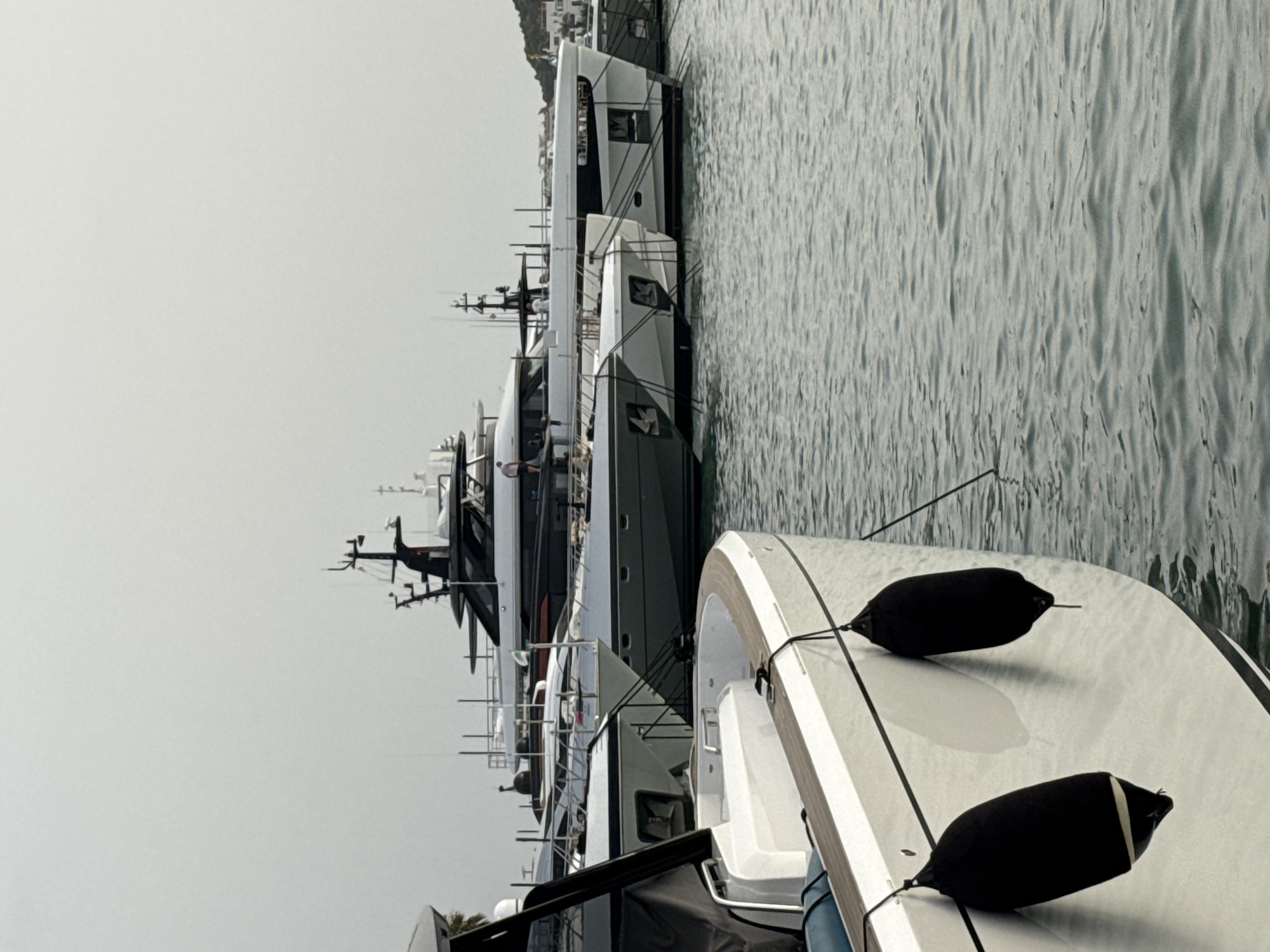 A fleet of yachts is docked in a harbor. In the foreground, the bow of a white yacht is visible, with two black fenders hanging from its side. In the background, several larger yachts are moored, displaying sleek, modern designs. The sky is overcast, and the water is calm and reflective. The scene suggests a luxury marina environment.