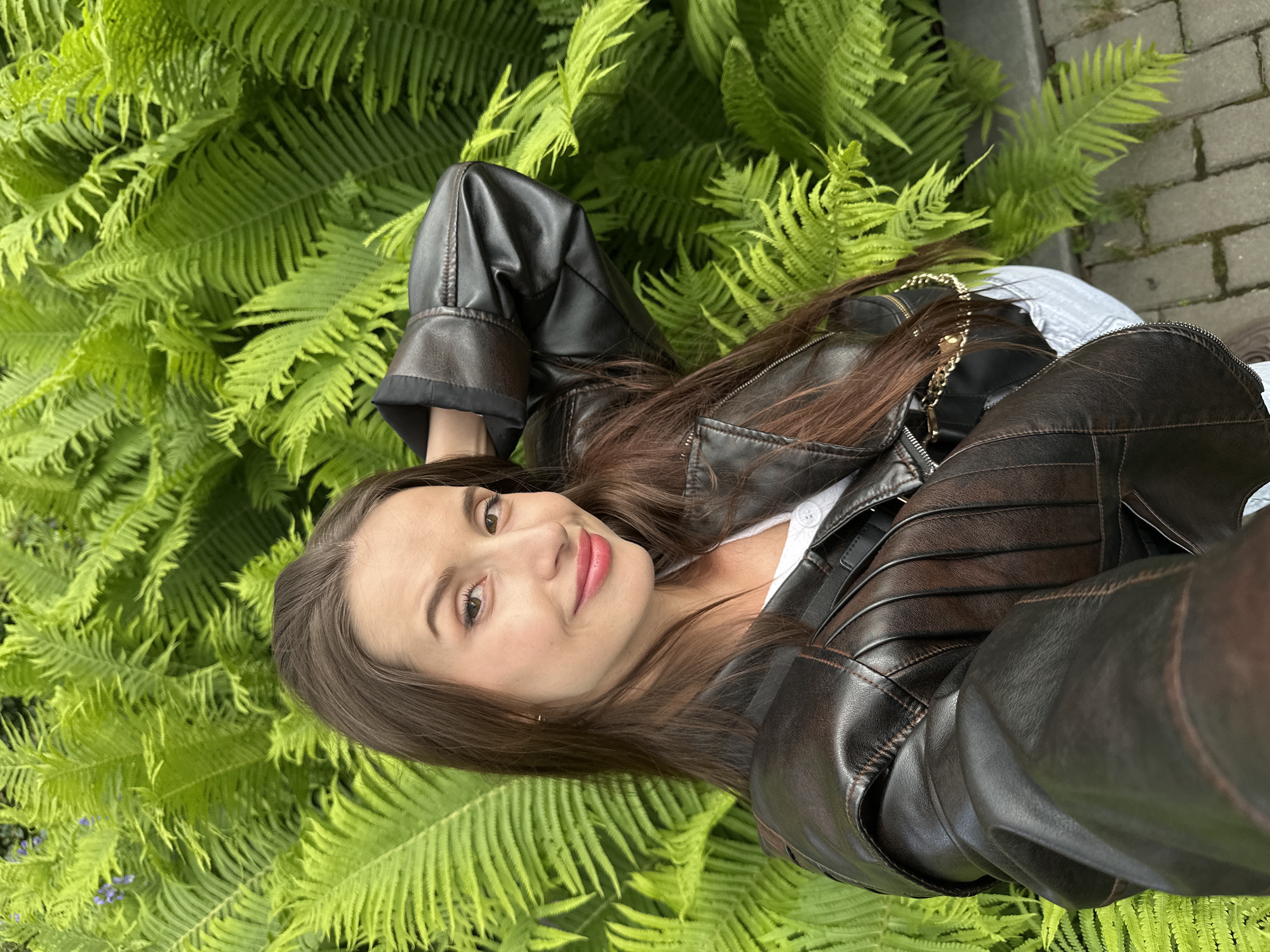 This rotated image captures a selfie of a young woman lying amidst lush, vibrant green ferns. She is looking directly at the camera with a gentle smile, exuding a relaxed and pleasant demeanor. Her long, dark brown hair is spread out around her head. She is wearing a dark brown leather jacket with visible stitching and quilted details on the sleeves and shoulders, over a white top. A gold-colored chain, likely from a bag, can be seen near her right shoulder. The background is almost entirely filled with the dense foliage of the ferns. On the far right edge of the image, a grey paved path, possibly made of bricks or stones, is visible, suggesting an outdoor setting like a park or garden in Yekaterinburg, Russia. The natural lighting indicates it is daytime. No text is visible in the image.