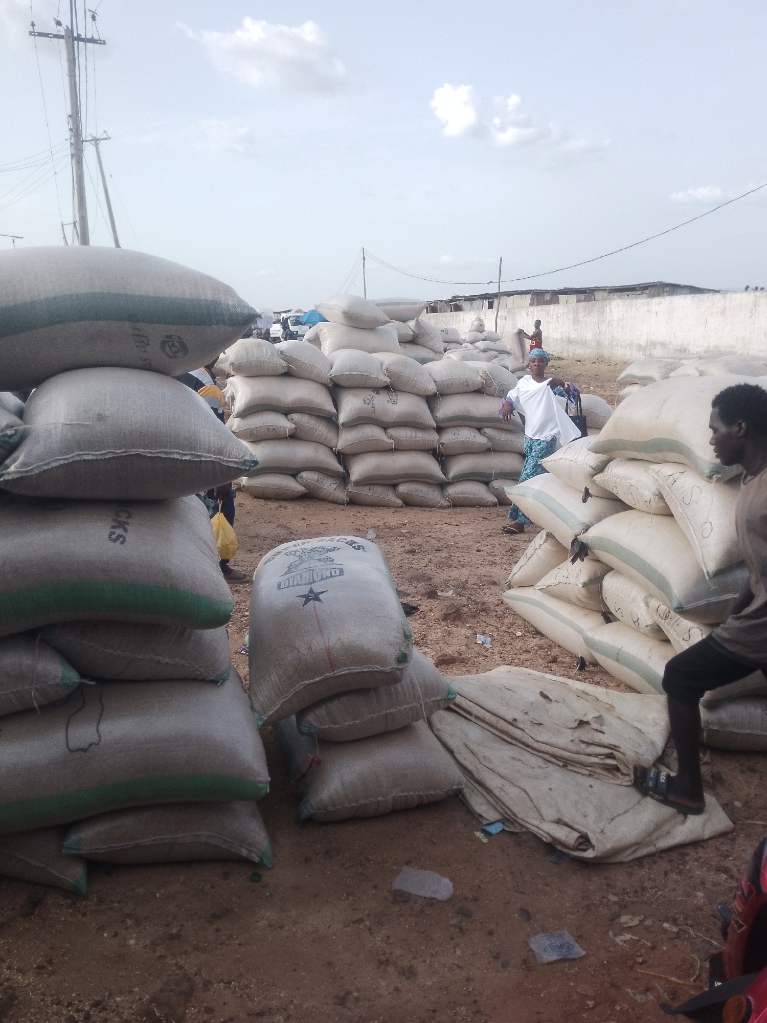 The image depicts a storage area with numerous stacks of burlap sacks in an open outdoor location. The ground appears to be dirt or gravel. The sky is overcast with some clouds. Several people are present, including a man standing near a stack of sacks on the right side of the frame, and a woman in a white top and blue patterned skirt standing near the center. A man with a red shirt is slightly visible behind the woman. In the background, there is a white wall and what appears to be a distant town or settlement. Power lines and poles are visible in the upper left corner of the frame. The location is in the city of Unknown, Unknown.