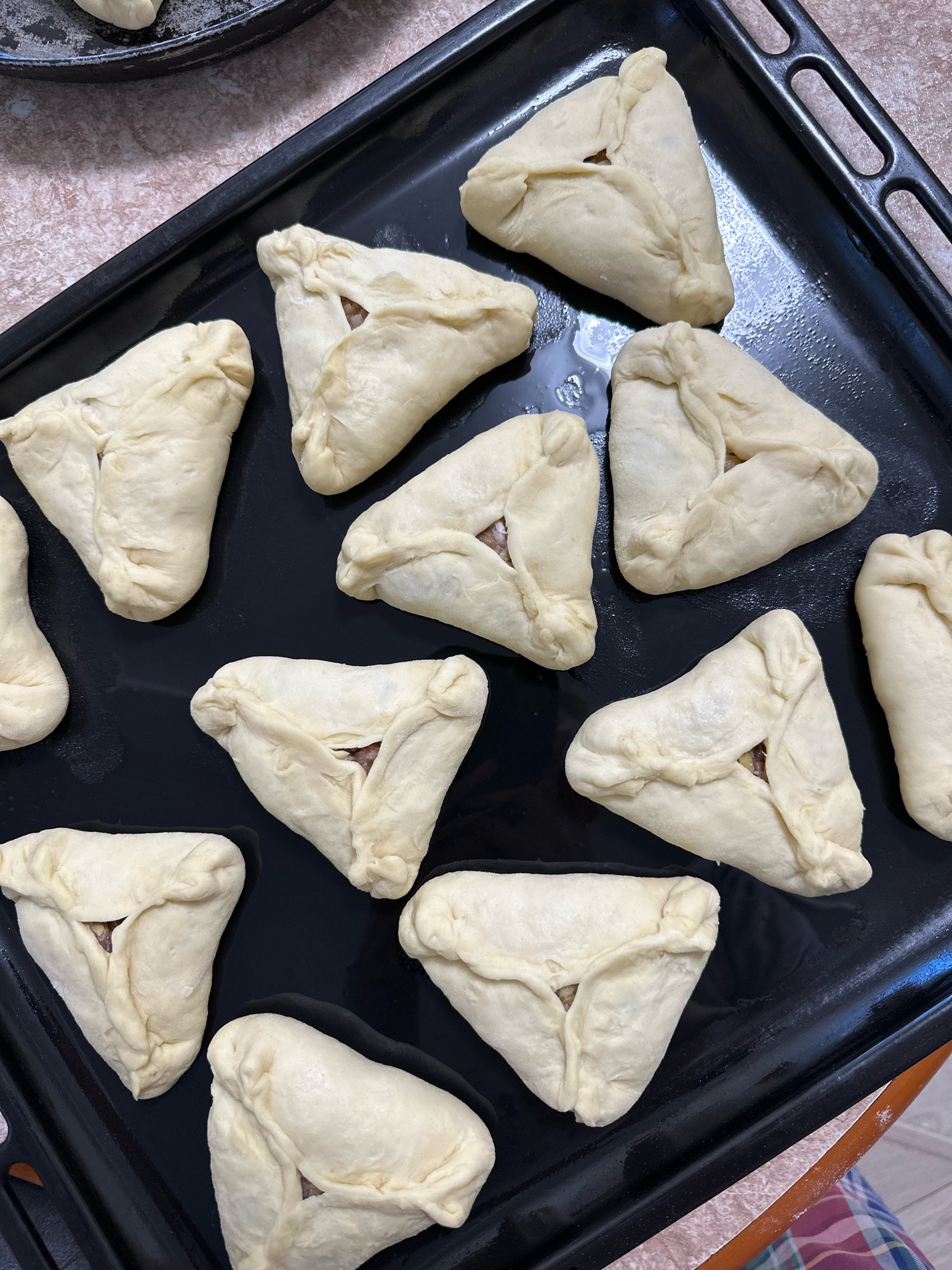 The image shows a close-up, overhead view of a black baking sheet filled with raw, triangular-shaped pastries, likely filled with meat, arranged on a textured countertop. The pastries are raw dough, folded to create a triangular shape with an opening in the center showing the filling. The baking sheet is on a light-colored, speckled surface. A portion of another baking sheet with pastries can be seen in the upper left corner, and the edge of a patterned fabric is visible at the bottom right. The scene is indoors, likely a kitchen, given the presence of baking equipment. The image does not contain any elements that would indicate a specific location in Buinsk, Russia.
