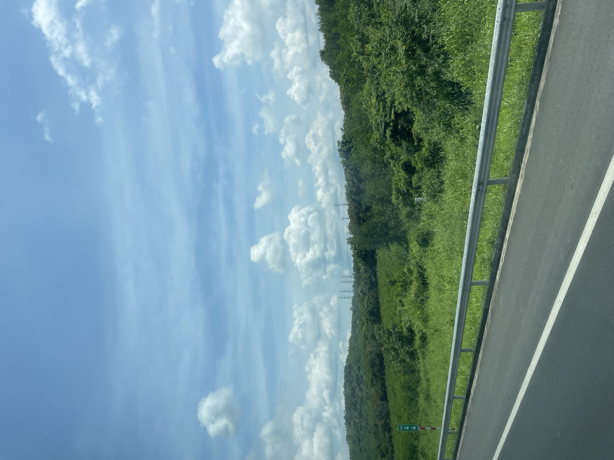 The image depicts a roadside scene in Tân Minh, Vietnam, on a clear day with a partly cloudy sky. In the foreground, a dark asphalt road with a white lane marker is visible. A silver metal guardrail separates the road from a grassy embankment. Beyond the embankment, lush green vegetation covers rolling hills. In the distance, several tall, lattice-style electrical transmission towers are visible against the horizon. A green signpost with white markings, likely a kilometer marker, is situated on the grassy shoulder near the guardrail. The overall impression is one of a rural or semi-rural highway setting.