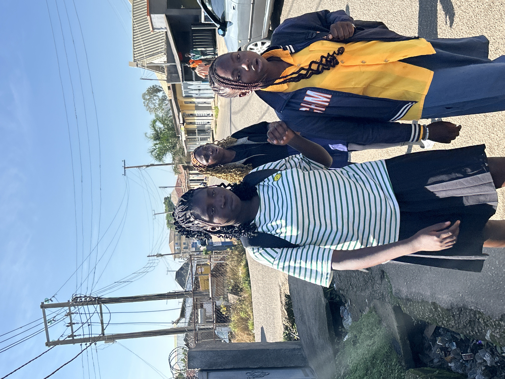 In an outdoor, urban setting in Jos, Nigeria, three young Black females, likely students, walk on a paved road. The foreground features two girls prominently, with a third slightly behind them. The girl on the left, closest to the camera, wears a green and white striped t-shirt and a black pleated skirt, carrying a backpack. To her right, another girl is dressed in a yellow button-down shirt, a dark jacket with white and orange trim, and a blue skirt, also carrying a backpack. The third girl, in the middle ground, has curly blonde hair and wears a black jacket over a dark top.
The environment suggests a commercial or residential area. Buildings with corrugated roofs and various storefronts line the street. Overhead, power lines crisscross the bright, clear blue sky, supported by wooden utility poles. A gray car is parked on the right side of the road. The road itself appears worn, and to the left, a concrete drainage channel is visible, containing some debris and overgrown vegetation. The overall scene depicts a common streetscape, possibly during daylight hours as indicated by the strong shadows.
