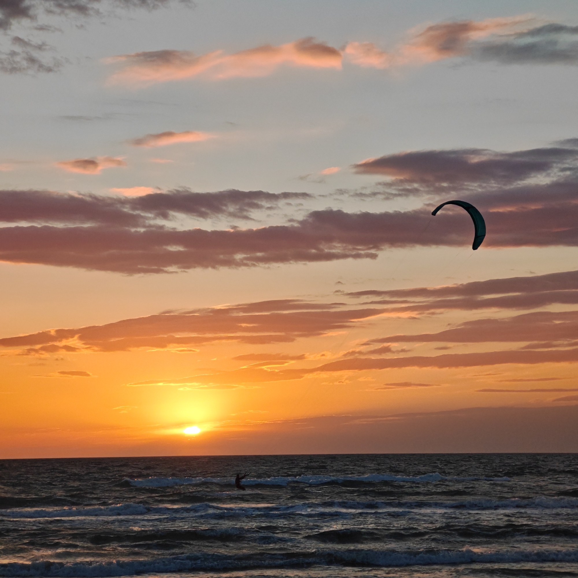 A wide-angle shot captures a sunset scene over the sea. The sun is setting on the horizon, casting an orange glow across the sky and reflecting on the water. Above the horizon are layers of clouds, colored in shades of orange, pink, and gray. On the water, there is a figure kitesurfing, with a dark kite visible in the sky. The waves are moderate and are closest to the viewer, receding towards the horizon. The photograph captures the scene in городское поселение Щелкино, Ukraine.