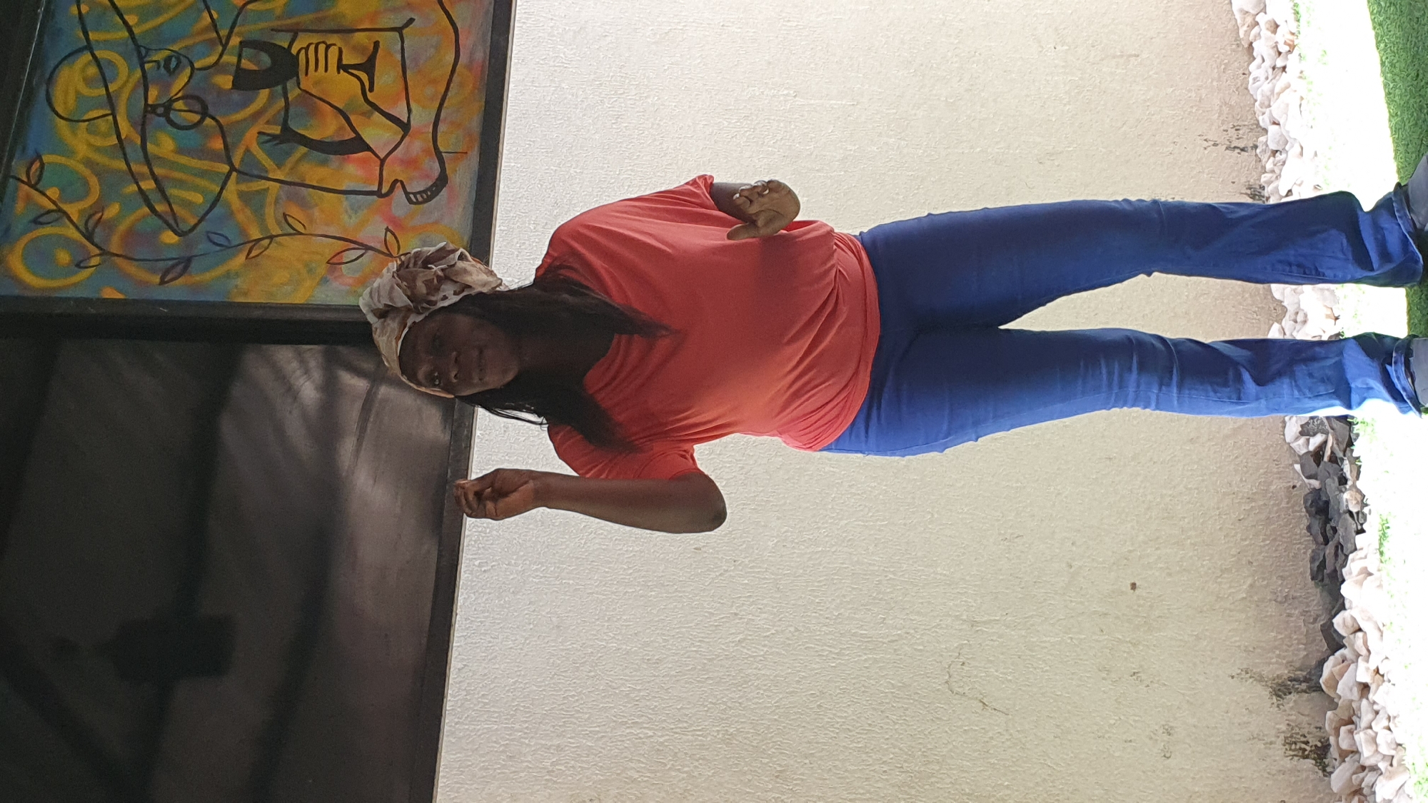 A medium shot captures a Black woman with dark hair and a patterned headscarf standing in front of a textured off-white wall. She is wearing a coral-colored, loose-fitting t-shirt with short sleeves and dark blue flared jeans. Her right hand is raised with her index finger pointing upwards, and her left hand is open, palm facing outwards. Behind her, a black framed abstract painting with yellow, blue, and red colors is visible, depicting a person wearing a wide-brimmed hat and holding a glass. The ground in front of the wall is covered with small white and dark stones and artificial green turf.