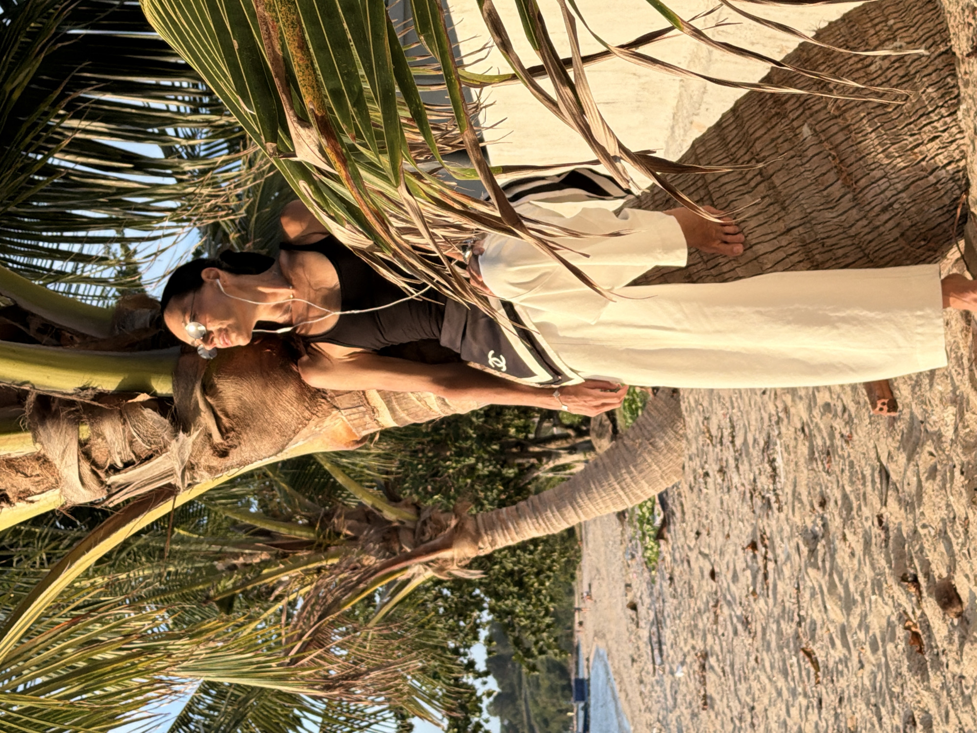 A woman wearing a black tank top, white pants, and round sunglasses is sitting on a palm tree on a sandy beach, in Ko Samui District, Thailand. She is leaning back against the trunk of the tree with her legs crossed. She is wearing earbuds and appears to be listening to music. The palm tree's fronds cast shadows on her and the surrounding sand. In the background, the ocean is visible, along with a line of trees and a small boat. The sky is clear and the sun is shining, creating a warm, golden light. The sand is light brown and appears to be a mix of fine grains and small shells. There is some debris scattered on the sand. The overall scene evokes a sense of relaxation and vacation.