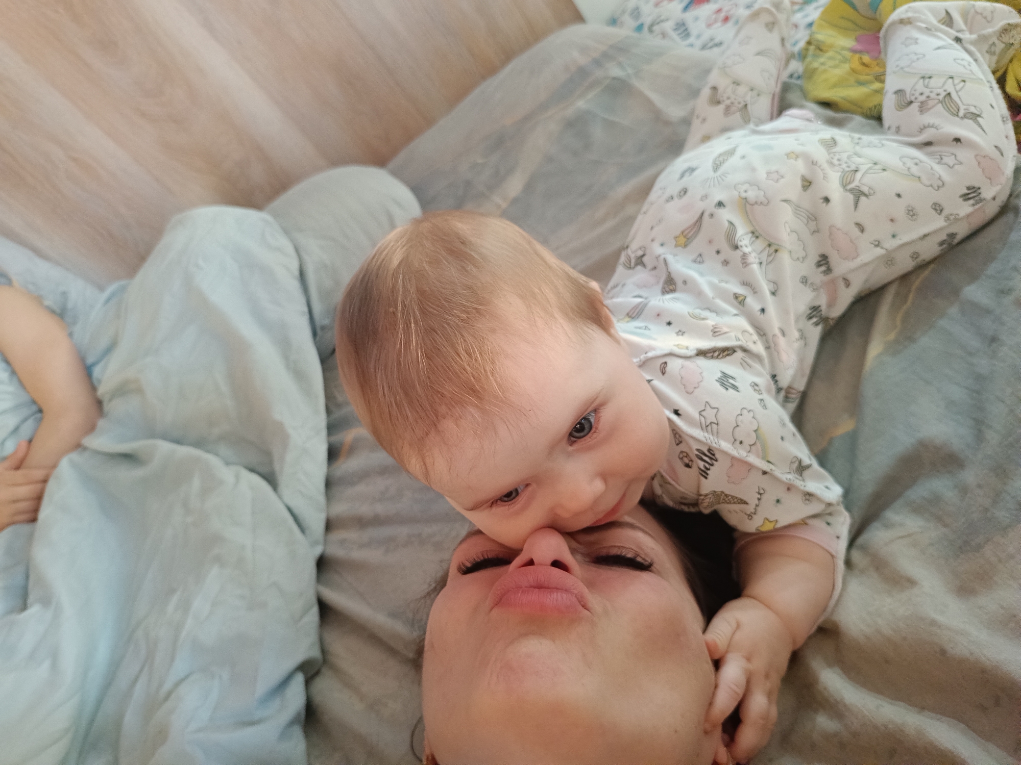 An overhead shot captures an intimate moment between a woman and a baby, lying on a bed or soft surface. The woman is on her back, facing the camera, with her head and upper chest visible. She has dark hair and is making a playful 'pouting' or 'kissing' expression with her lips.
The baby, with light reddish-blonde hair and blue eyes, is positioned perpendicularly above the woman, resting its head near her face. The baby's nose or mouth is gently touching the woman's nose. It wears a white sleepsuit adorned with colorful patterns, including unicorns, clouds, stars, and the legible text "hello sweet." The baby's body extends away from the woman, with its left arm bent and hand near the woman's neck.
The setting appears to be indoors, likely a bedroom. Various soft fabrics are visible: a light blue blanket or duvet to the left, a greyish sheet or blanket beneath both individuals, and a patterned yellow fabric further in the background behind the baby. A wooden floor is partially visible in the top left corner, suggesting a home environment. The scene conveys a moment of affection and playfulness between the two. The location context provided is Rusalevka, Russia.