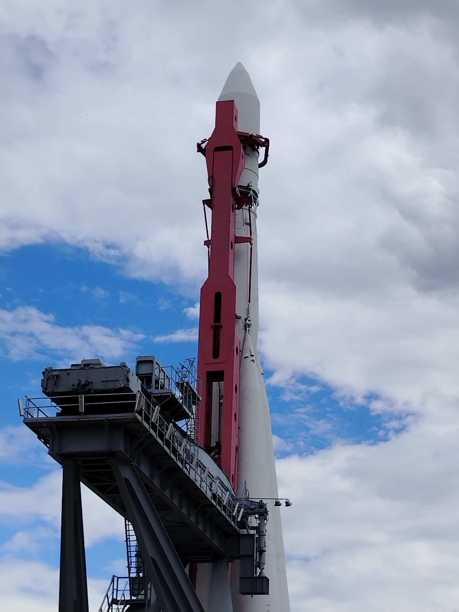 This low-angle, outdoor, medium shot captures a large white rocket with a red pylon structure attached, standing vertically against a sky with scattered clouds. The rocket is situated on a complex metal launch platform, which is supported by large, angled metal beams. The platform features multiple levels with railings and some visible machinery. The rocket's nose cone is white and pointed, and the main body is also white, with segments visible along its length. The red pylon is a prominent feature, extending along the side of the rocket and appearing to support it. The sky is a mix of blue patches and large, fluffy white and grey clouds.
The rocket and launch platform are located at the Museum of Cosmonautics in Moscow, Russia. There are no people visible in the image.