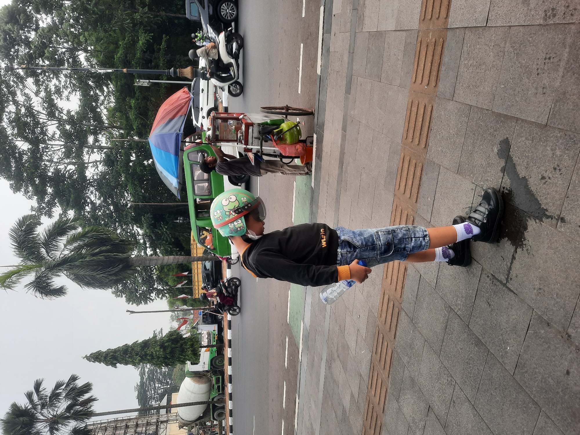 In Sukaraja, Indonesia, a young boy wearing a helmet, black hoodie, and denim shorts stands on a sidewalk next to a street. He holds a partially consumed bottle of water in his right hand. The sidewalk is paved with square stone tiles and has tactile paving for the visually impaired.
Adjacent to the sidewalk, a street bustles with activity. A food cart, covered by a colorful umbrella, is attended by a man. He is seen interacting with the cart, which has a green gas cylinder attached. Several vehicles are present on the road, including a cement mixer truck, a green van, a white scooter with a rider, and a black car. Motorcyclists are also visible in the background.
The environment suggests a moderately overcast day, with lush green trees forming a backdrop to the urban scene. The presence of palm trees adds a tropical feel to the location. The road features white lane markings.