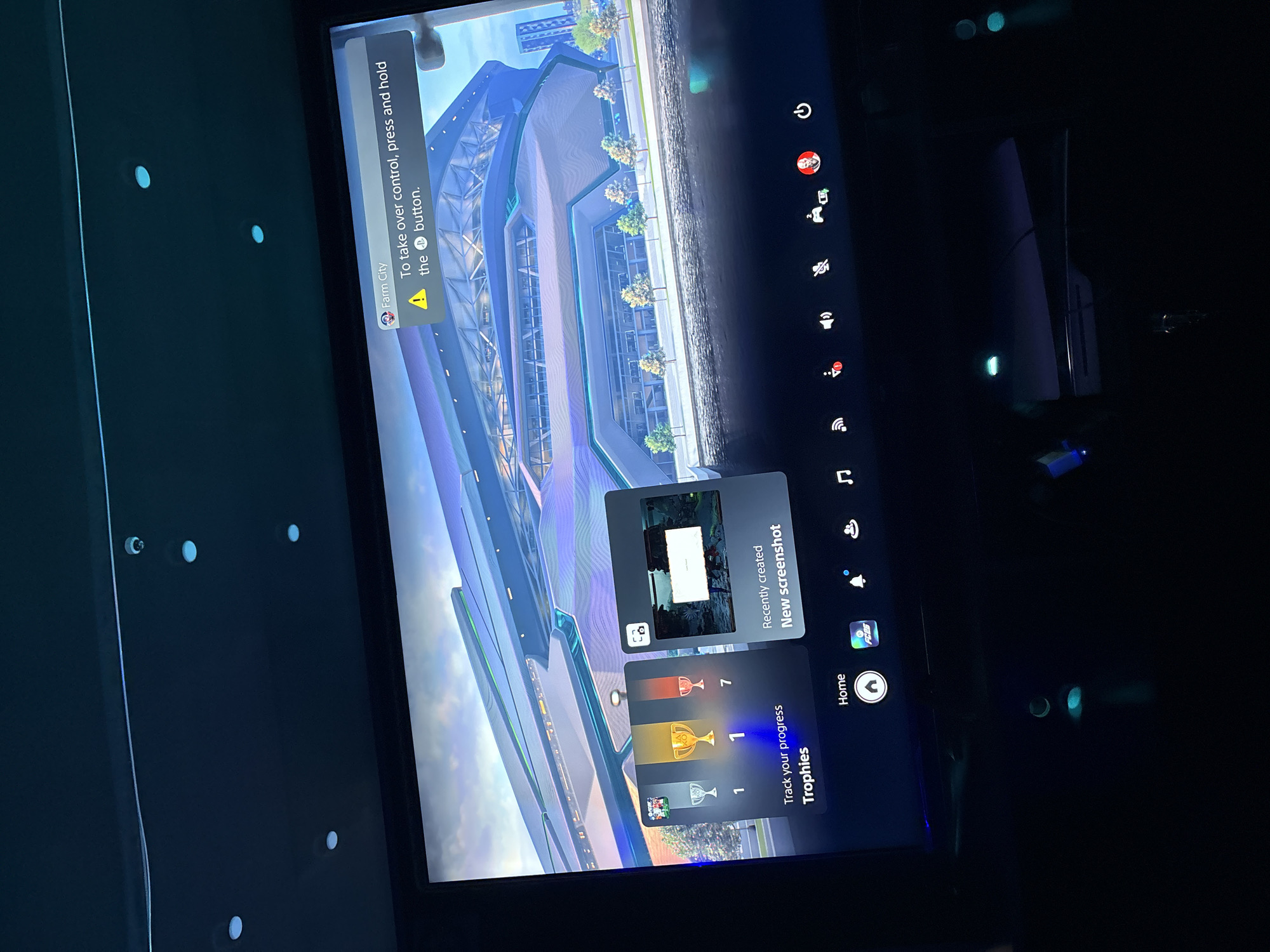 The image shows a television screen displaying a video game interface, likely on a PlayStation console. The game appears to be a city-building or simulation type, with a complex architectural structure visible on the screen. The interface includes notifications and trophy information. The PlayStation interface is visible on the right side of the screen. The environment surrounding the screen appears dark, with some LED-type lighting visible on the left.
The image does not provide any information related to Abuja, Nigeria.