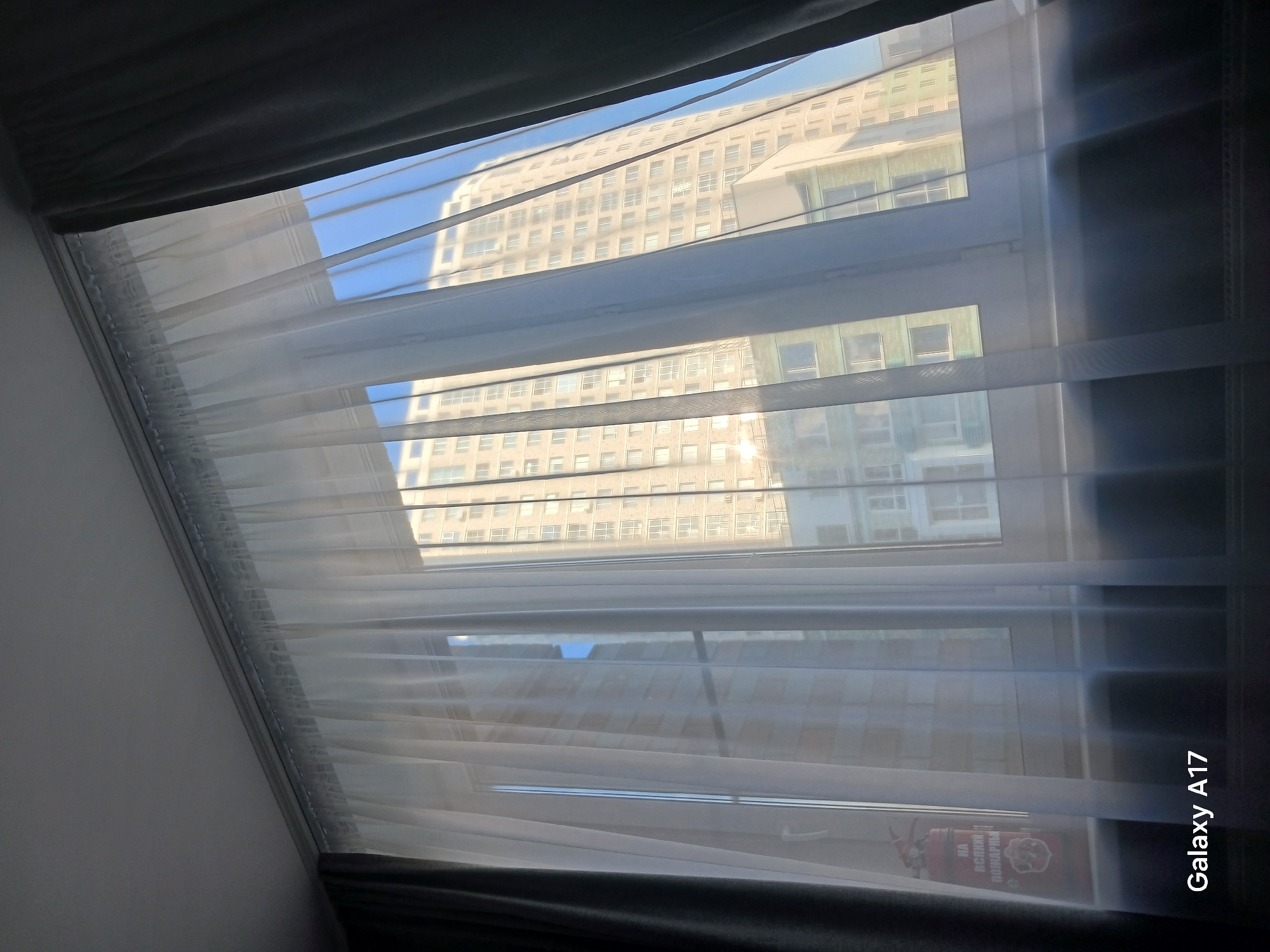 The view through a window, presumably in an apartment in Yekaterinburg, Russia, is partially obscured by a sheer white curtain. Outside, a multi-story building with a light-colored facade and rows of windows dominates the scene. The sky is visible in some areas, suggesting a daytime setting. In the foreground, a dark-colored curtain or drape frames the view. A sticker is visible on the bottom right part of the window. The "Galaxy A17" is printed on the bottom right of the image.
