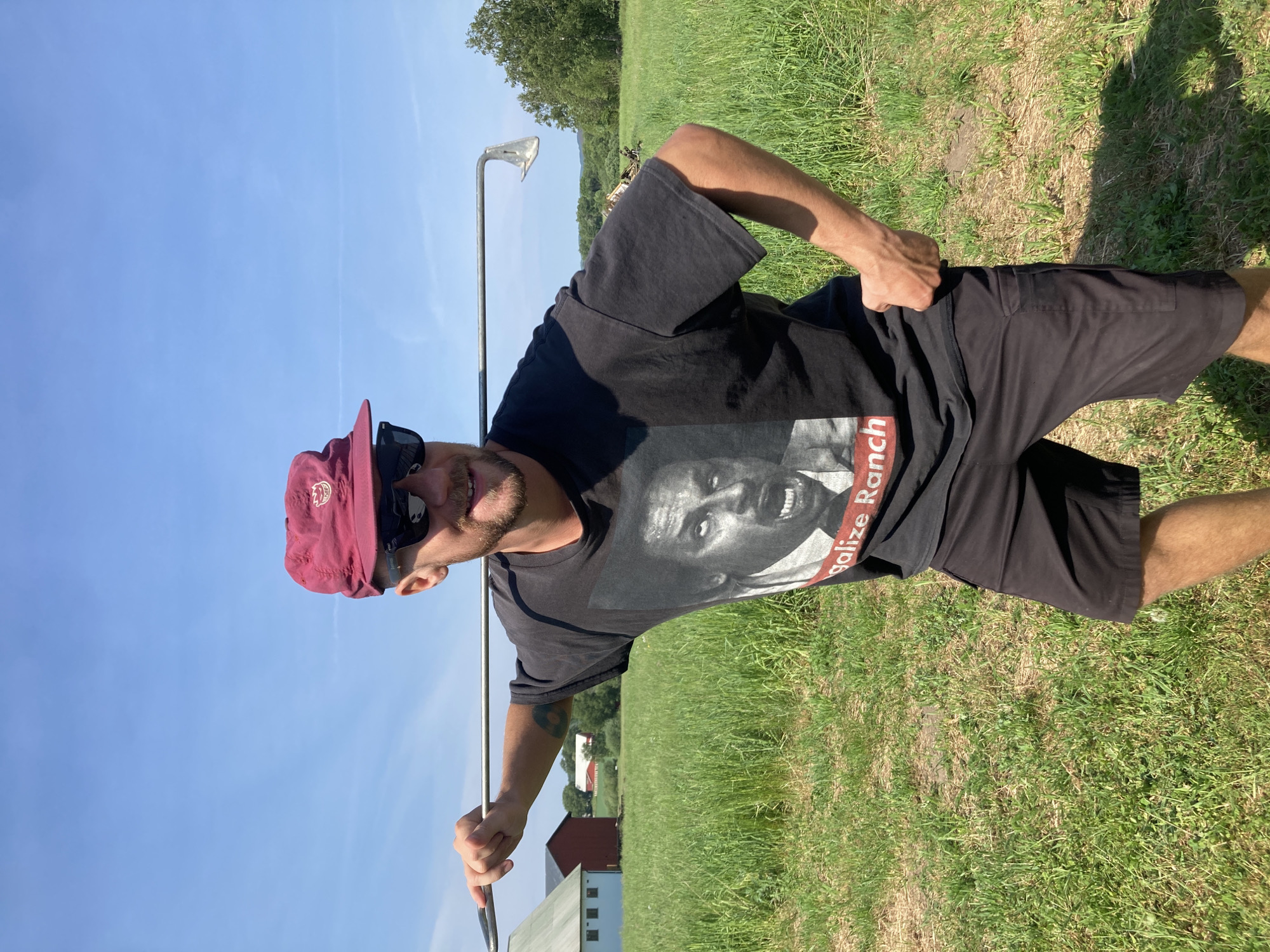 A man is depicted outdoors, smiling broadly and looking directly at the camera on a bright, sunny day. He is wearing a black t-shirt, black shorts, and a red baseball cap with sunglasses perched on top. He has facial hair, appearing as stubble or a short beard. His right hand holds a long, thin metal object, resembling a golf club or practice swing aid, pointed towards the ground. His left hand is placed on his hip.
The man's black t-shirt features a large grayscale graphic of a person's face, seemingly with an open mouth or expressing themselves, possibly related to speech or singing. Below this graphic, red text is visible, partially reading "galize Ran" or "galize Ranch" (likely "Legalize Ranch").
The setting is an open, grassy field or meadow with varying shades of green grass. In the background, lush green trees line the horizon, and beyond them, several red farm buildings, including what appears to be a barn, are visible under a clear, bright blue sky. The scene suggests a relaxed moment in a rural, agricultural landscape, consistent with a location like Vessigebro, Sweden. The time of day is clearly daytime, given the bright sunlight.