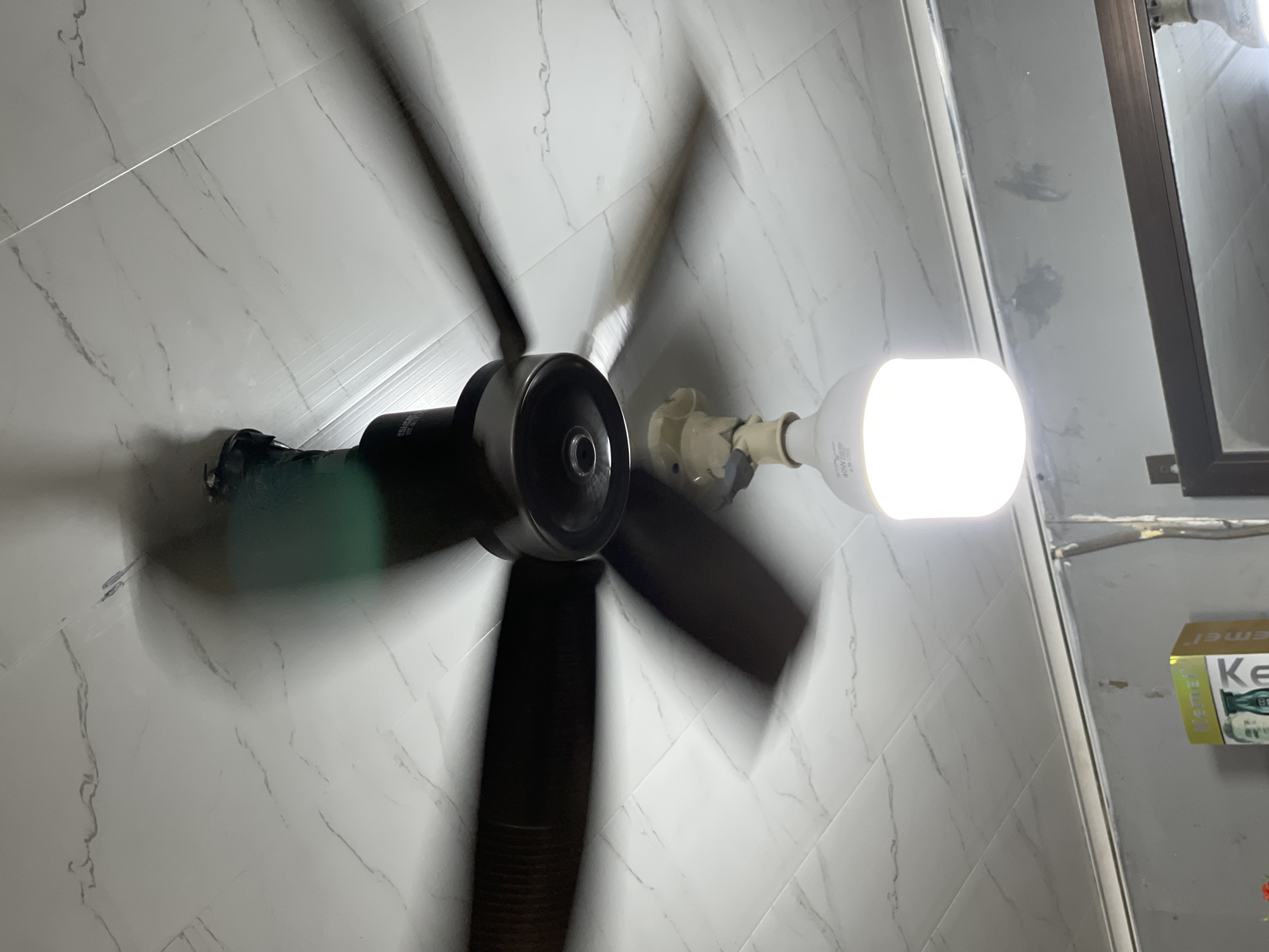 The image shows a ceiling fan with dark-colored blades spinning in front of a white tiled ceiling. A bright LED light bulb hangs from the center of the fan. The lighting and motion blur suggest the image was taken indoors, likely in a residential or commercial space. There are no people visible in the image. The overall scene appears to be an ordinary interior view of a room. The location information about Cempeudak, Indonesia is external to the visual content of the image and cannot be inferred from it.