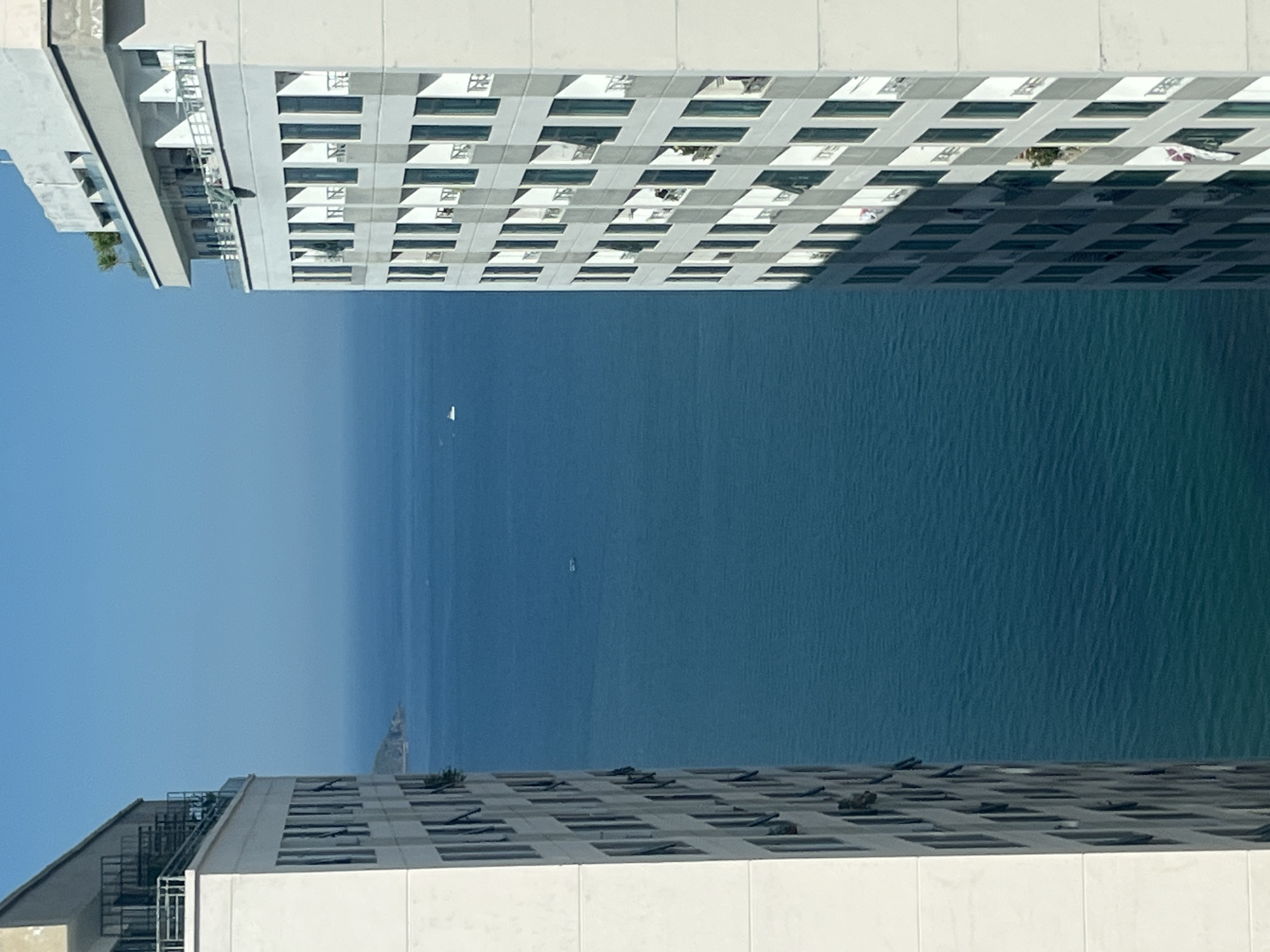 The image presents an upward view between two tall, modern buildings in Bac Nha Trang Ward, Vietnam, revealing a clear blue ocean stretching to the horizon. The buildings are predominantly white, featuring a grid-like pattern of windows and balconies. The sea is calm with a few distant sailboats visible. The sky transitions from a deep blue to a lighter hue near the horizon. A rocky point of land is visible in the distance.