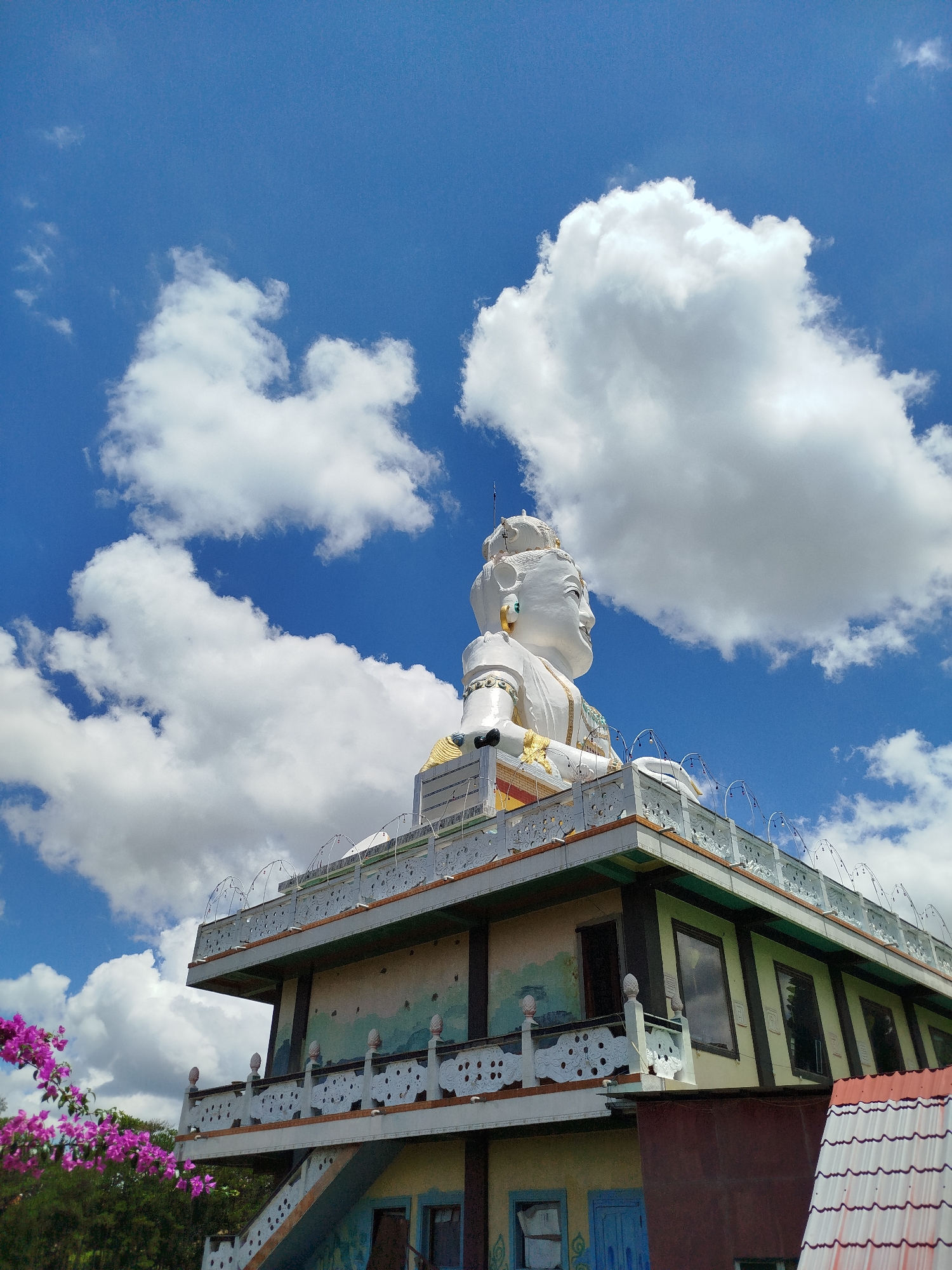 A low-angle, eye-level shot depicts the elevated statue of Lady Buddha at Linh Phuoc Pagoda, a temple in Da Lat, Vietnam. The white statue is positioned atop a multi-tiered building, its head nearly merging with the backdrop of a vibrant blue sky dotted with cumulus clouds. A portion of the pagoda's structure is visible, including balconies, windows, and partial views of its decorative elements. In the lower left corner, pink bougainvillea flowers add a pop of color against the green foliage.