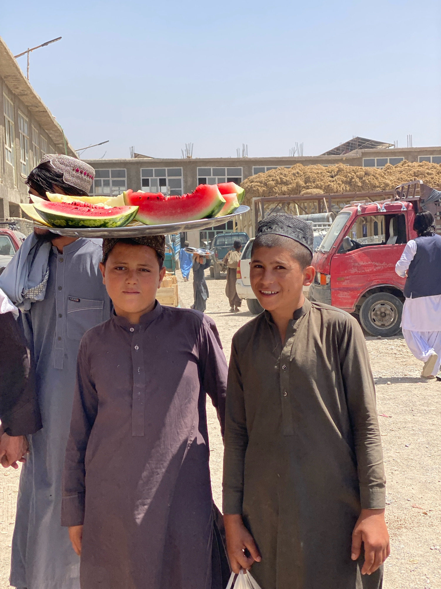 Two young boys, likely teenagers, are the primary subjects in the foreground of this outdoor scene, which appears to be a marketplace in the city of Kandahar, Afghanistan. The boy on the right is smiling at the camera, wearing a dark green tunic and a black cap. He is holding a white plastic bag in his right hand. The boy on the left is facing forward with a neutral expression, also wearing a dark tunic and a patterned cap.
Behind the boys, a man is partially visible, carrying a large metal platter on his head that holds several sliced watermelons and a piece of melon. He is wearing a gray tunic and a traditional patterned cap. The background reveals a bustling environment with elements of a city market. A red truck is parked on the right, and other vehicles and structures are visible, including buildings under construction with exposed rebar. Piles of hay or straw are stacked high behind some of the structures. The ground is dusty and appears to be unpaved. The overall lighting suggests a bright, sunny day.