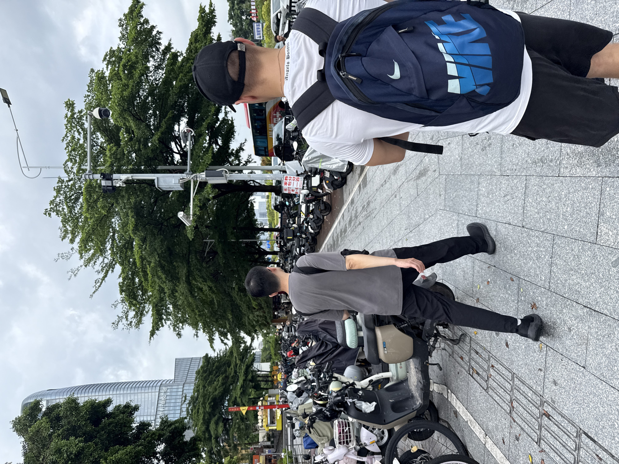 The image depicts a street scene in Haizhu District, China. Two individuals are the primary subjects. The first, on the left, is a person wearing a grey t-shirt and black pants, seated on an electric scooter. They are looking towards the right. The second person, on the right and slightly behind, is wearing a white t-shirt, black shorts, and a black baseball cap. They have a navy blue backpack with a large Nike logo on their back. This person is looking downwards and to the left.
The scene is outdoors, with a paved sidewalk and a street lined with numerous parked electric scooters. Lush green trees are visible on the left side of the frame, with a glimpse of a modern, glass-fronted building in the background. Overhead, the sky is overcast, suggesting a cloudy or possibly rainy day. Traffic lights and street lamps are also present, indicating an urban environment.
The overall mood appears casual and everyday, with no strong emotional indicators. The lighting suggests it is daytime, likely late morning or afternoon, due to the diffused light.
Visible text includes the Nike logo on the backpack and Chinese characters on some of the signs and scooter bodies, though they are not clearly legible. One sign near the top features Chinese text, and there is also a yellow sign with red characters visible further down the street.