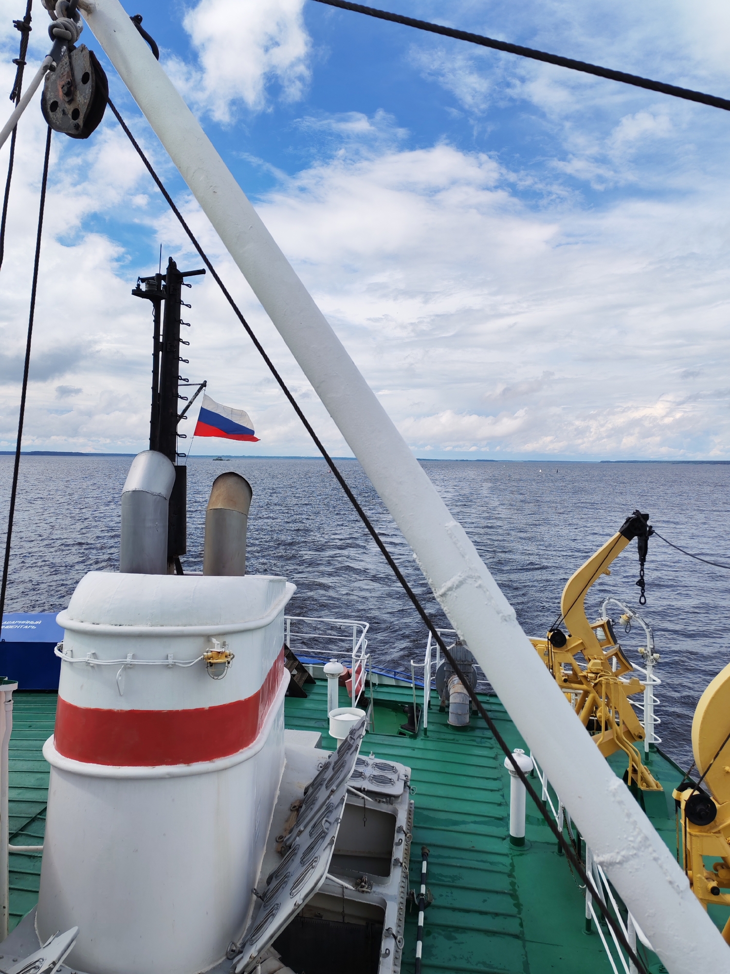 An eye-level, medium shot captures a view from the deck of a vessel on a body of water under a cloudy sky. In the foreground, a large white pipe bisects the frame diagonally from left to right. To the left, a white and red smokestack dominates the mid-ground, with a Russian flag waving from a pole just behind it. A yellow crane is visible to the right of the smokestack. The green deck of the ship extends into the background, meeting the dark blue water. The horizon line is visible in the distance, with a faint outline of land under a sky filled with white and gray clouds. The image appears to be taken in or near Kostyaevka, Russia, given the presence of the Russian flag.