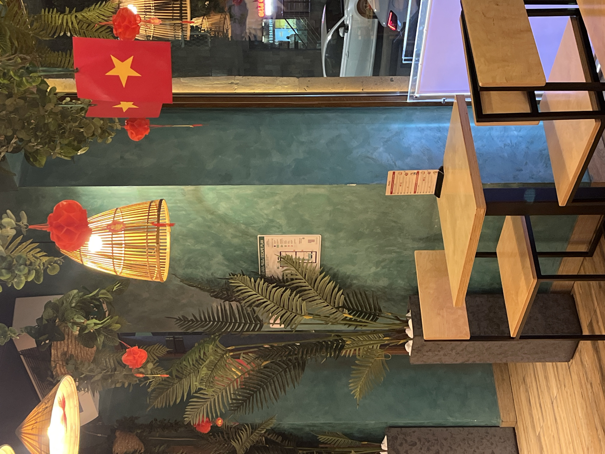 The image depicts the interior of a restaurant in Yerevan, Armenia. Hanging from the ceiling are decorative Vietnamese flags and bamboo lanterns. A modern shelving unit with wooden panels is on the right. Artificial plants, including palm fronds, are visible. A menu holder is attached to one of the shelves. The walls have a textured green finish. A reflection of the outside street can be seen in a window above the flags.