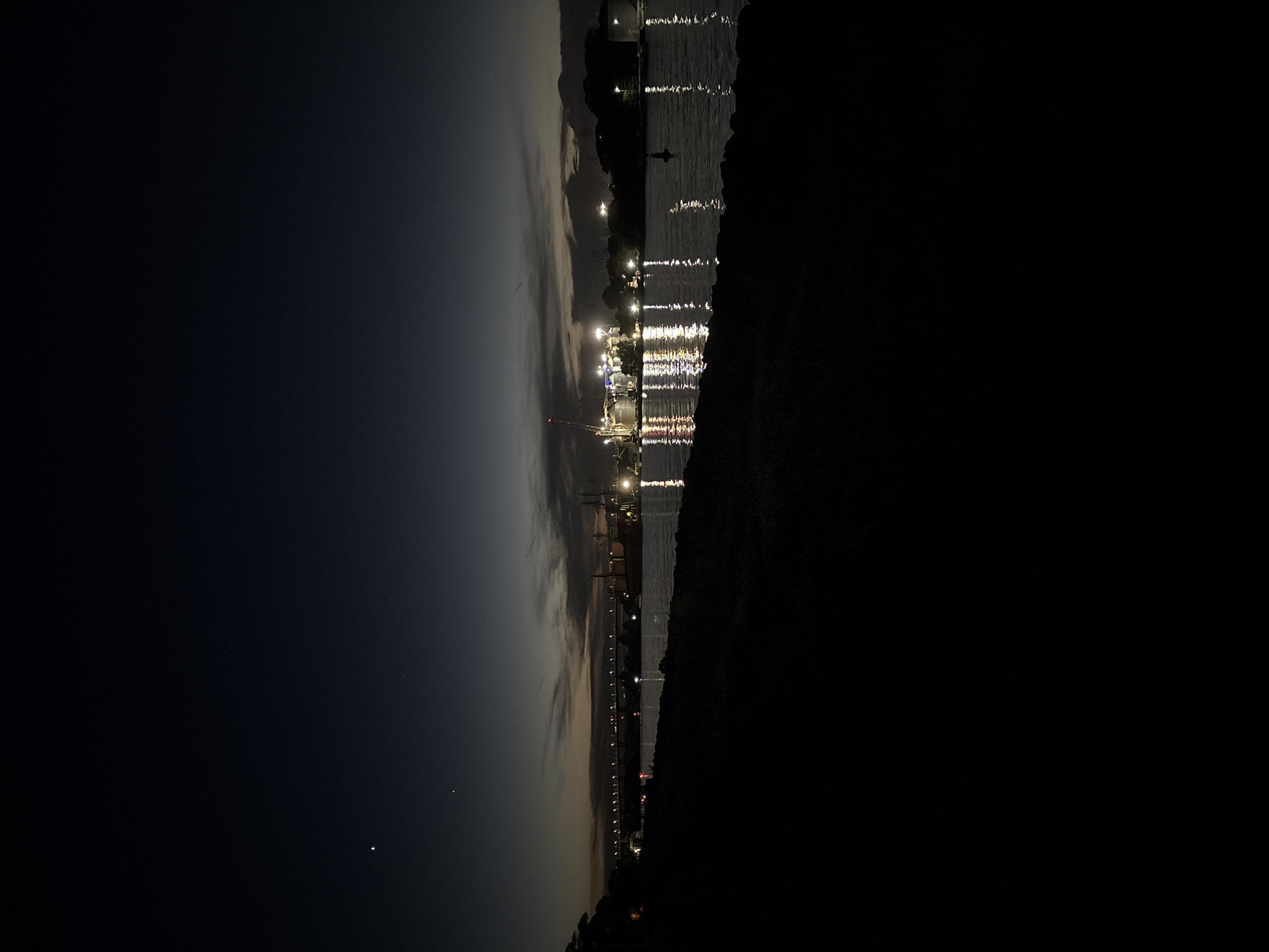 The image depicts a waterfront scene at dusk or night in Varna, Bulgaria, with artificial lights reflecting on the water. In the distance, a long bridge is illuminated by a series of lights. Further along the waterfront, a port area is visible with industrial structures, cranes, and lights from buildings, possibly silos. A large ship is docked near the port. The foreground shows a dark, silhouetted embankment covered in vegetation. The sky is a deep blue with some faint clouds, and a few stars or celestial bodies are visible in the upper left quadrant. The overall scene suggests a view of the Varna port and the Asparuhovo bridge, a significant landmark in the city. There are no people or animals visible in the image.