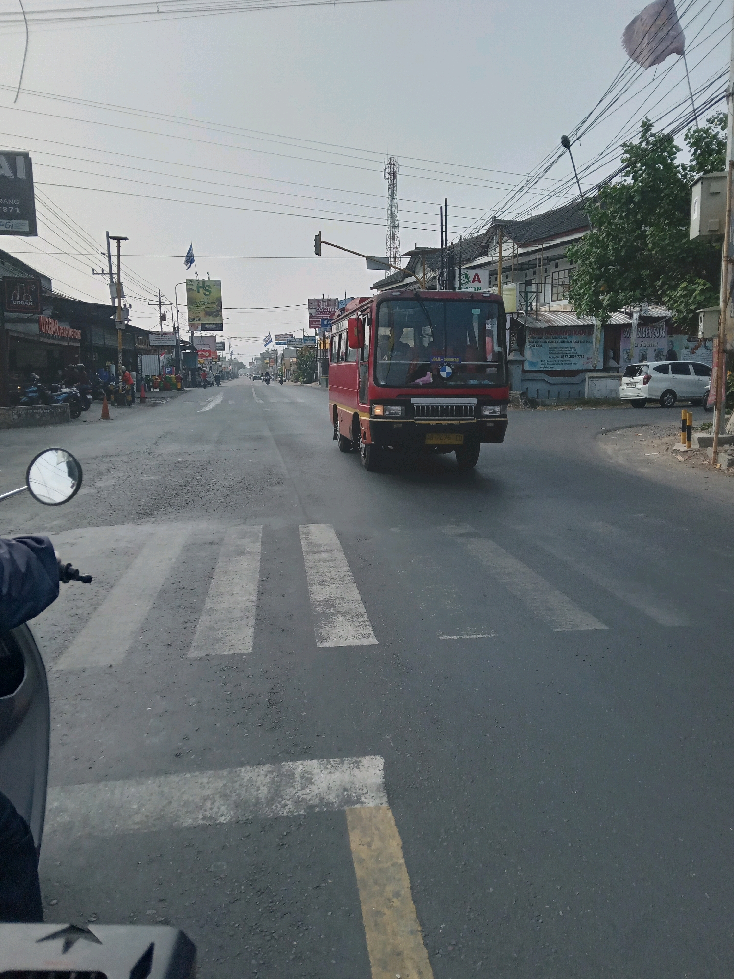 The image shows a street scene in Banguntapan, Indonesia, on a slightly overcast day. In the foreground, a motorcycle's rearview mirror and handlebars are visible from the perspective of the rider, indicating the photo was taken from a moving motorcycle. The road is paved and has a pedestrian crossing marked with white stripes. A red bus with the number plate "AB 7475 CD" is traveling on the right side of the road, approaching the pedestrian crossing. On the left side of the road, there are several shops and buildings, with motorcycles parked in front of some of them. Several flags are flying on poles along the street, including one tattered white flag. A telecommunication tower is visible in the distance. The overall environment suggests a bustling commercial and residential area in an Indonesian city.