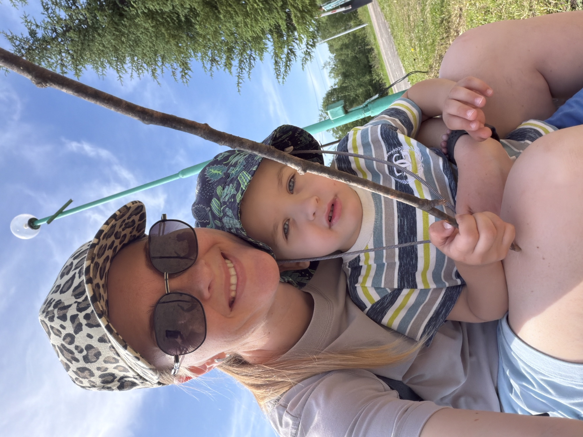 A woman and a young child are in the foreground of this medium shot, with the woman on the left and the child on her lap. The woman is wearing a leopard print baseball cap and sunglasses, and a light-colored t-shirt. She is smiling and looking at the camera. The child is wearing a bucket hat and a striped t-shirt. He is holding a stick and looking away from the camera. Behind them, there is a blue sky with some clouds, a green tree, and a lamppost. The setting appears to be outdoors, possibly a park or a playground, in Kul'dur, Russia. The overall mood of the image is relaxed and happy.