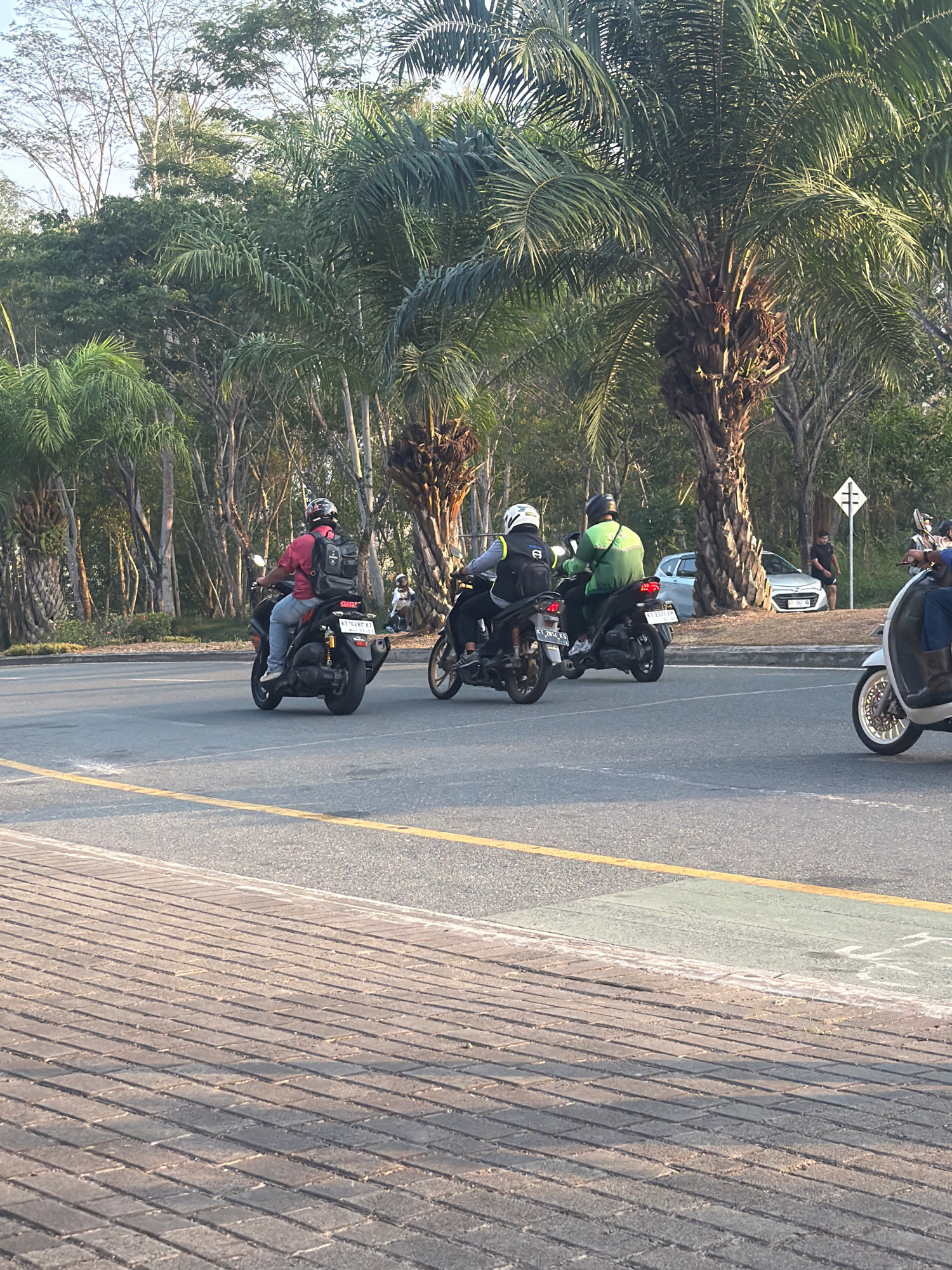 The image depicts a street scene in Balikpapan, Indonesia. In the foreground, a paved walkway with rectangular bricks runs parallel to a road. The road features a yellow lane divider and a green painted section on the right side. Several motorcycles are in motion on the road. One motorcycle on the left carries a rider in a red shirt and a black backpack, wearing a helmet. To its right, another motorcycle is ridden by a person in a green jacket and helmet, also carrying a backpack. Further to the right, a white scooter with a rider in dark clothing is partially visible. In the background, lush green trees and palm trees line the street, creating a tropical ambiance. A silver car is parked on the side of the road, and a traffic sign with intersecting lines is visible. Several people are seen standing or walking in the distance. The lighting suggests it is daytime, possibly late afternoon, with soft sunlight casting shadows.