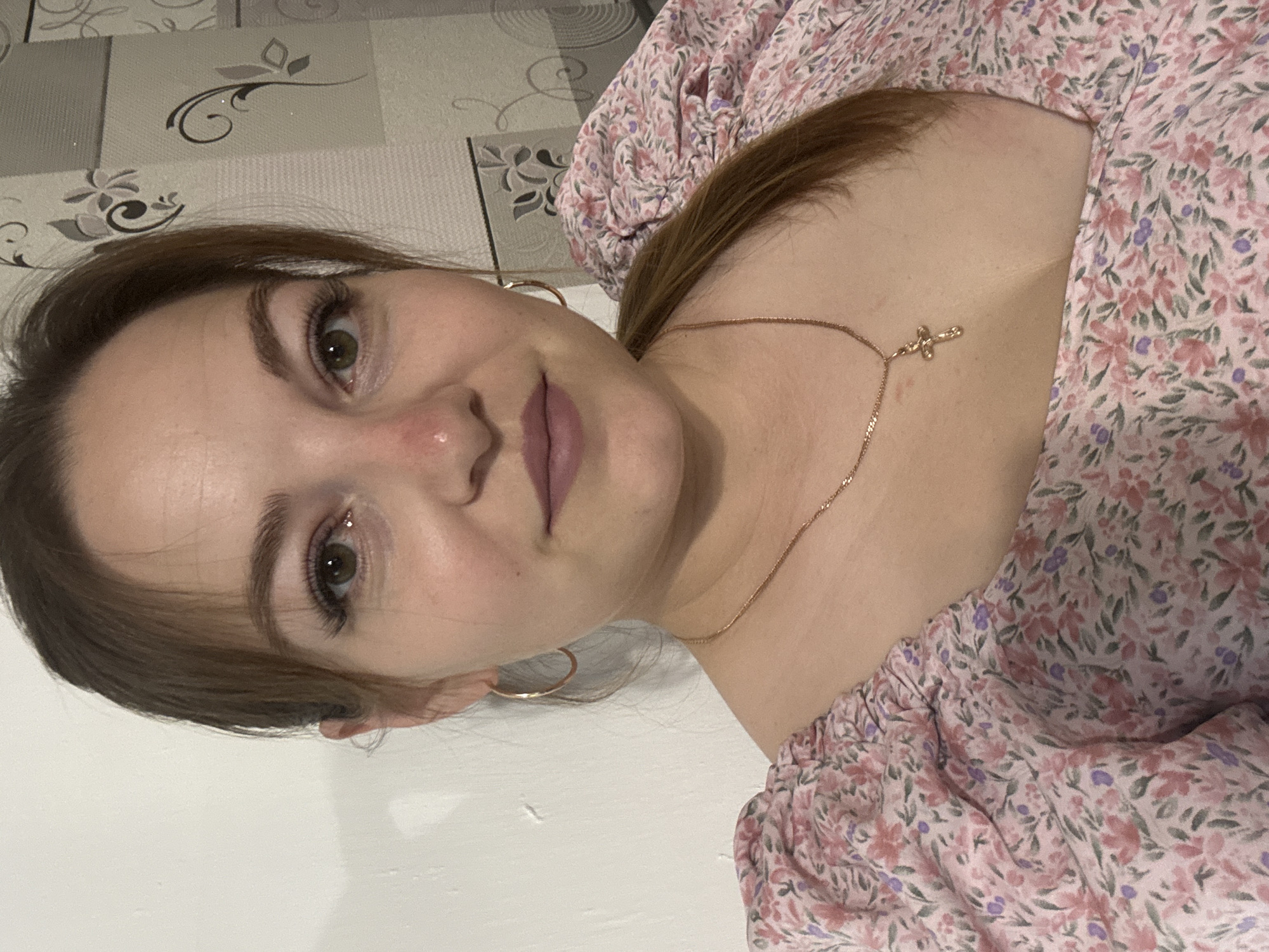 A medium shot photo of a woman looking at the camera. She is wearing a pink floral print dress, a gold chain necklace with a cross pendant, and gold hoop earrings. Her hair is pulled back into a ponytail. The background appears to be a wall with a floral wallpaper. The image does not provide any information about the location in the city of Zubov, Russia.