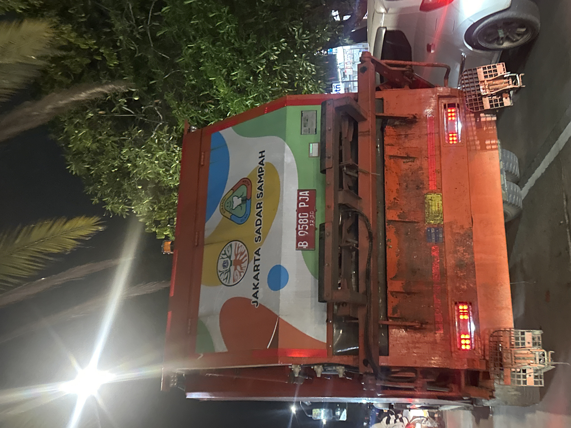 The image captures the rear of an orange and red garbage truck with a colorful design on its back, which reads "JAKARTA SADAR SAMPAH" (Jakarta Aware of Garbage). The license plate "B 9580 PJA" is visible, and the design indicates the vehicle is likely part of Jakarta's municipal waste management fleet. The truck is positioned on a road at night, with other vehicles nearby, including a white car partially visible above the truck. Trees and streetlights are also visible. The license plate suggests the location is likely in Jakarta, Indonesia. Given the presence of streetlights, trees, and other vehicles, it is likely the image was taken on a street within the city of North Jakarta.
