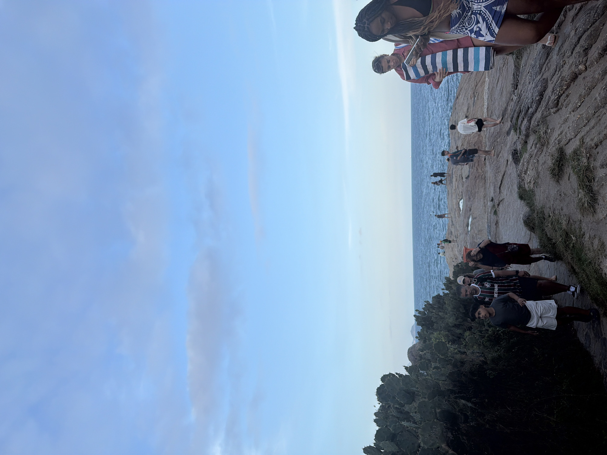 This image depicts a group of people walking along a rocky coastline with lush vegetation on the left and the ocean stretching out to the horizon under a pale blue sky with scattered clouds. In the foreground, two women are visible, one in a black bikini top and a blue and white patterned skirt, and the other in a red and white striped shirt, holding a phone and a towel. To their left, a family is walking away from the camera; the boy in front wears a gray t-shirt and white shorts, the man behind him wears a striped soccer jersey with "BETANO" written on it, and a woman behind him wears a dark shirt and skirt. Further in the distance, several other individuals are scattered along the rocky path and near the water's edge. The rocky terrain suggests a coastal path, likely in a park or a natural reserve. Based on the visual cues, such as the rugged coastline, the type of vegetation (Opuntia cactus), and the warm climate implied by the attire of the people, this scene is likely located at the Arpoador Rock, a popular scenic spot in Rio de Janeiro, Brazil, known for its stunning sunset views and rocky landscape overlooking Ipanema and Copacabana beaches.