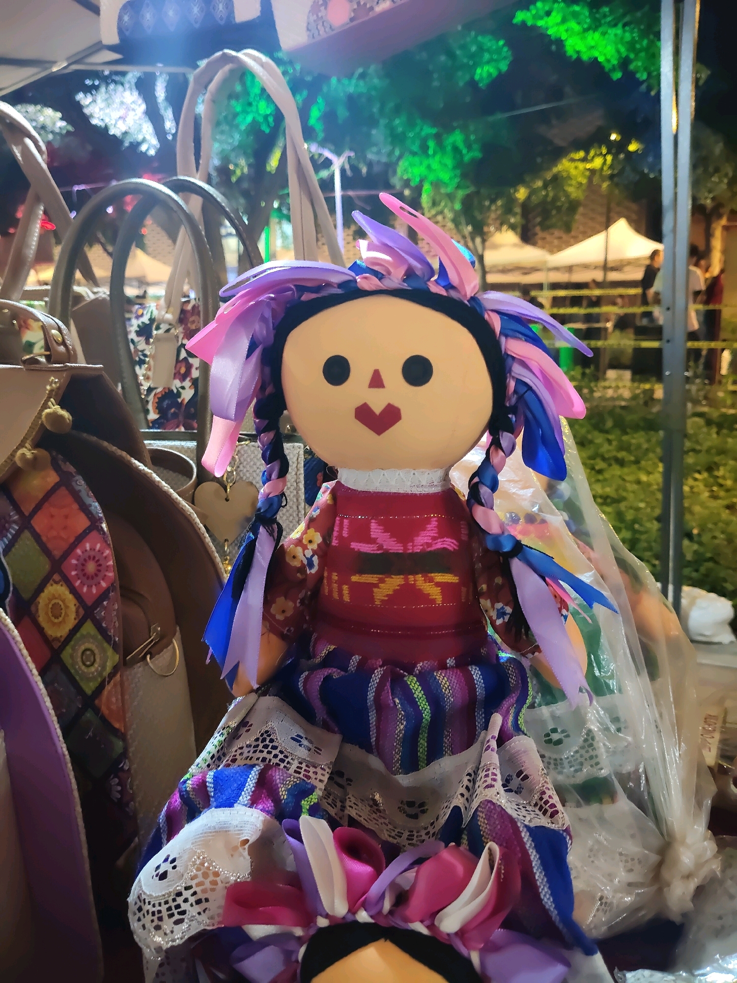 This is a vertical, medium shot, outdoor, nighttime photograph of a large, traditional Mexican doll, known as a "Lele" doll, displayed in a market setting. The doll is the central subject, facing forward. It has a round, tan face with black dot eyes and a red heart-shaped mouth. Its hair is styled with colorful ribbons in shades of pink, blue, and purple, braided into two pigtails. The doll wears a vibrant red shirt adorned with colorful woven patterns in yellow, green, and blue. Its skirt is made of layered, colorful fabrics, including striped material in purple, blue, and white, and a white lace trim.
The doll is positioned among various market items, including several large tan handbags with visible handles and clasps, and other decorative fabrics with geometric patterns. The background is blurred, suggesting a busy market environment. In the distance, there are white tent structures, green trees illuminated by artificial lights, and the silhouettes of people. The lighting indicates a festive or commercial event, possibly a night market. The overall scene conveys a sense of cultural display and commerce. Without further information or context, it is not possible to definitively pinpoint the exact location within Hermosillo, Mexico.