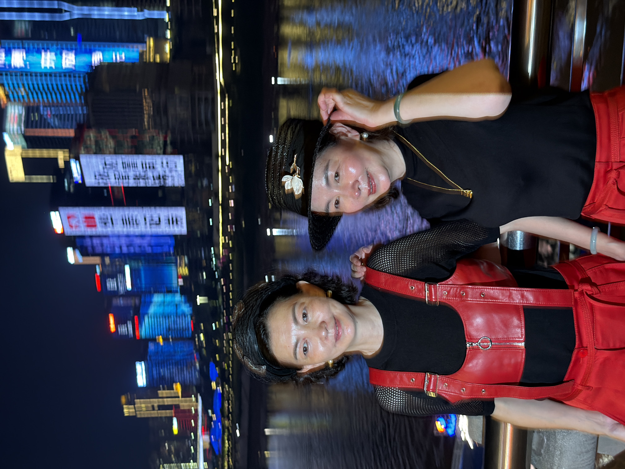 Two women are posing for a photo at night in front of a body of water with a city skyline in the background. The woman on the left is wearing a black top with a red leather harness over it, and red pants. She has short dark hair and is looking towards the camera. The woman on the right is wearing a black top, red pants, and a black hat with a floral embellishment. She has short dark hair and is also looking towards the camera, adjusting her hat with her right hand. The city skyline consists of tall buildings illuminated with colorful lights. The scene appears to be along a waterfront promenade in Yuzhong District, China, likely Chongqing, given the distinctive skyline.