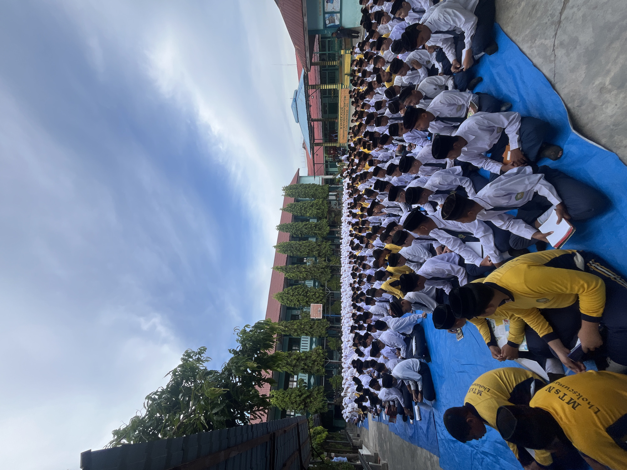 An aerial perspective captures a large group of male students, all wearing white shirts, dark pants, and black peci hats, sitting in rows on a bright blue tarpaulin spread across a paved outdoor area. The students are facing the same direction, suggesting they are attending an assembly or event. Some students hold books or papers. In the background, a two-story building with green and red accents and numerous windows stands, partially obscured by tall, conical trees. Further in the background, another building with a red roof and light blue walls is visible. The sky above is partly cloudy with a mix of white clouds against a pale blue backdrop. The scene appears to be taking place in the school grounds of MTsN Lhokseumawe, given the lettering visible on the yellow shirts of some students in the foreground, which reads "MTsN Lhokseumawe."