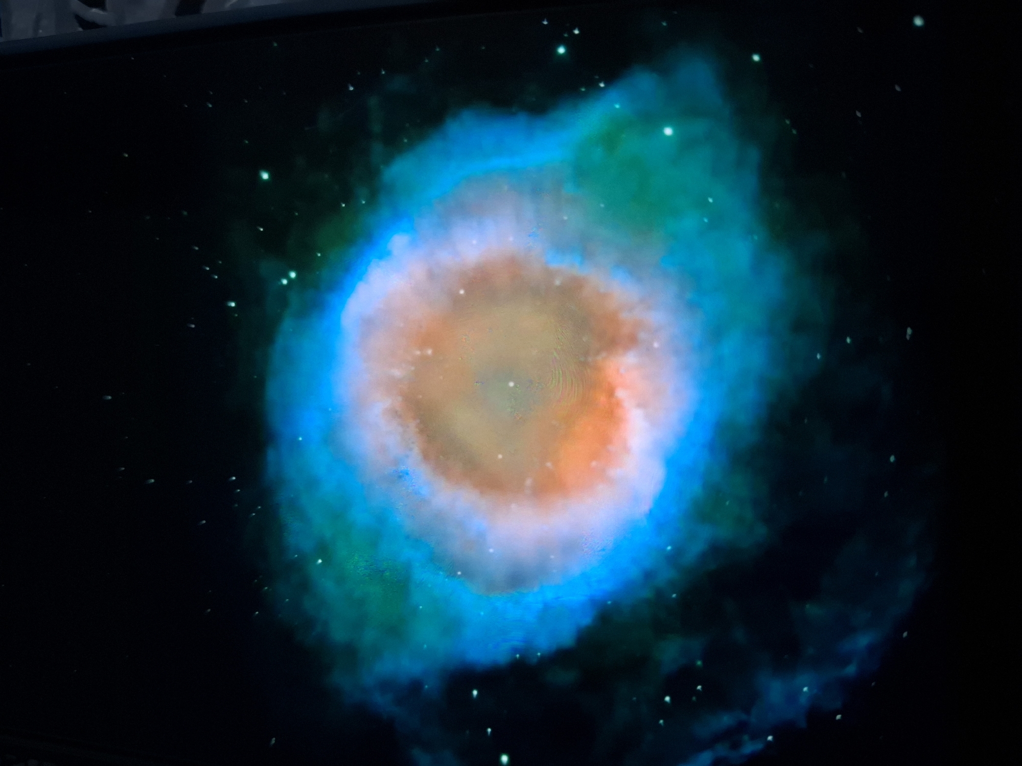 The image displays a colorful nebula in outer space. The nebula is predominantly blue and green, with a central area that appears orange and yellow. Numerous small white dots, representing stars, are scattered throughout the dark background. The image is framed by a dark border on the left and right edges, and a light gray border on the right. The scene is not located in a city.