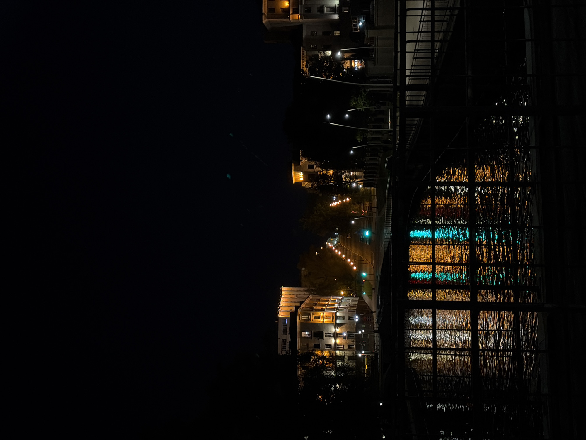 The image shows a night scene in an urban environment. Buildings are illuminated by streetlights and internal lights, with reflections visible in a body of water. A grid-like structure, possibly a bridge or railing, is in the foreground, partially obscuring the view. Streetlights line a road that appears to lead towards the horizon. The overall impression is one of an urban nightscape with a focus on light and reflection. The location is Unknown, Unknown.