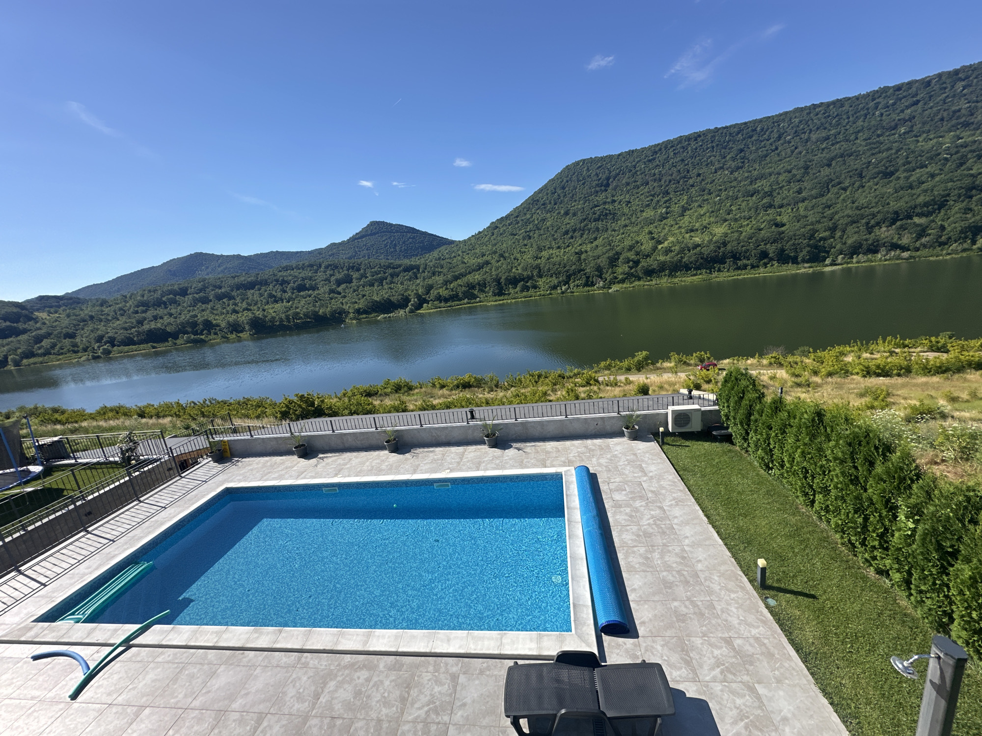An outdoor swimming pool area is situated in front of a body of water, backed by lush, tree-covered hills under a clear blue sky. The pool, rectangular in shape, is filled with bright blue water and covered with a rolled-up blue solar cover on one side. The poolside is paved with large, light-colored square tiles. A green pool hose lies on the tiles near the edge of the pool. In the background, a dark metal fence borders the property, beyond which is a grassy area with a few small trees. A row of tall, dense green hedges runs along the right side of the image, adjacent to a well-manicured lawn. Two black lounge chairs are placed on the tiled area. In the distance, beyond the pool and lawn, is a wide, dark green body of water reflecting the sky, and beyond that, rolling hills covered in dense green foliage. The overall scene depicts a serene and scenic resort or private residence setting.