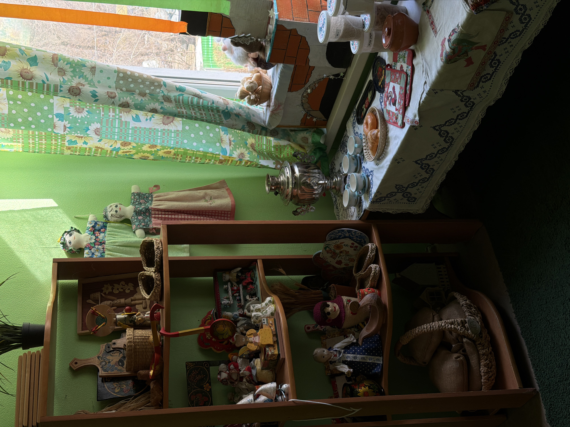 A cluttered interior scene. The location is impossible to ascertain based on the given image. A wooden shelf unit houses dolls, straw goods, and other folk objects. Above the shelves, there is a samovar on a tablecloth with cups and food. A window is visible, draped with patterned fabrics. There is also a light-colored tablecloth, possibly on a table, holding jars and kitchen linens. The overall color scheme is dominated by greens, browns, and whites. The room appears to be domestic, filled with objects suggesting craft or folk art. The lighting appears to be somewhat low. The objects appear to be arranged on a shelf.