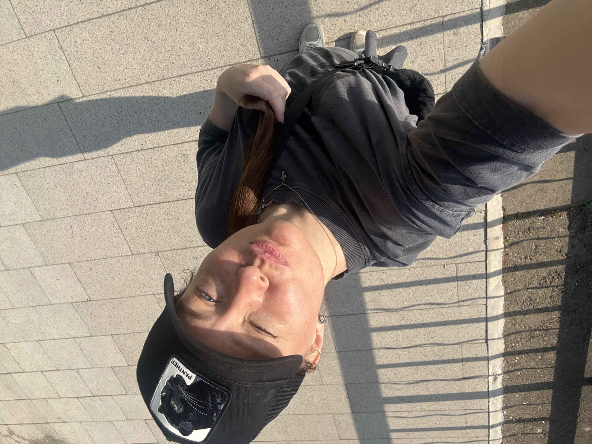 A person, seen from a high angle, stands on a brick-paved surface. The person is wearing a black cap with a "Panther" patch, a gray sweatshirt, and gray shoes. The person is holding their hair with one hand. A shadow falls across the brick pavement. The location is possibly Chaikovsky, Russia.