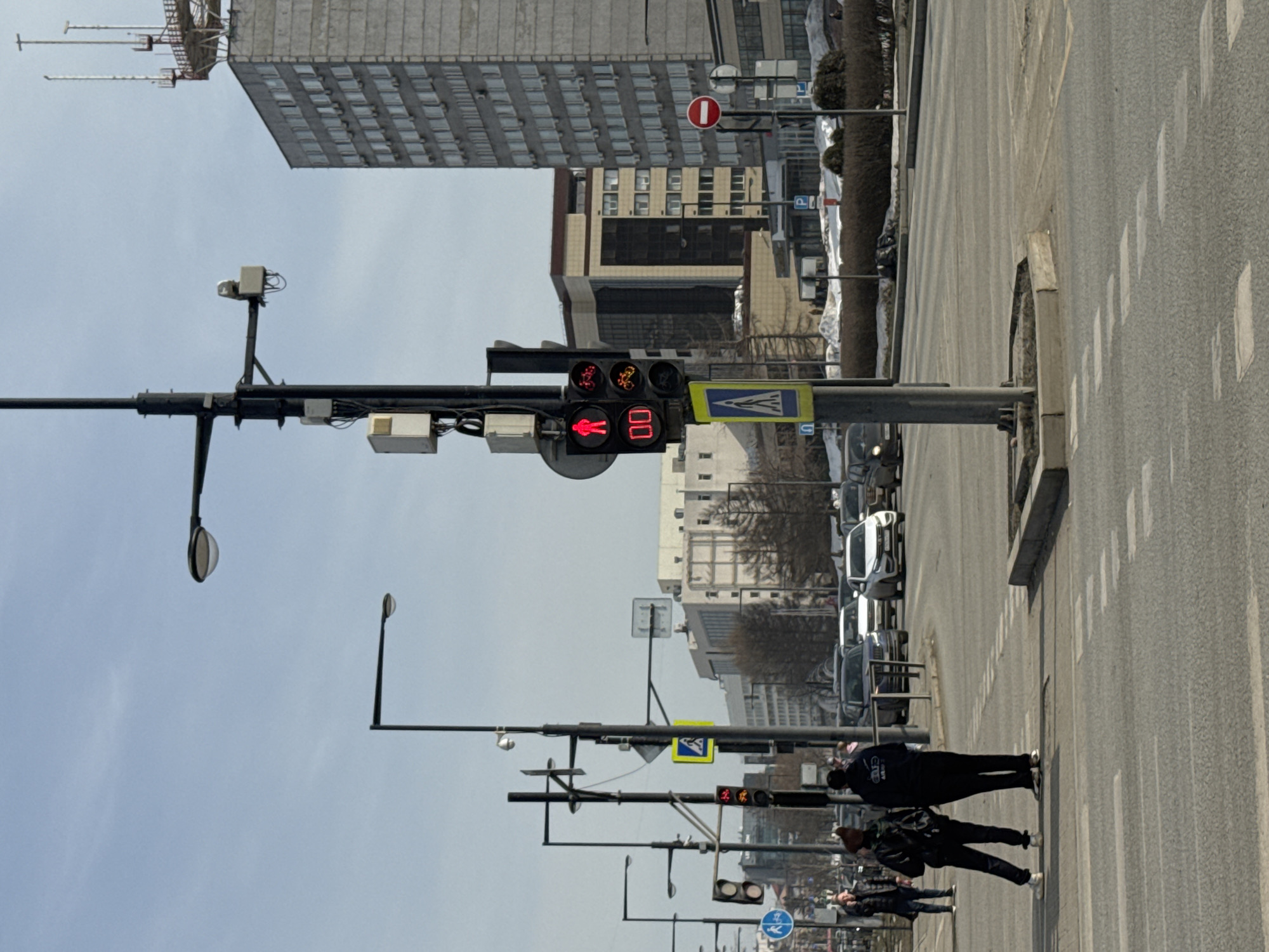 The image captures an urban street scene in Perm, Russia, specifically at or near the intersection of Komsomolsky Prospekt and Ulitsa Lenina. The prominent tall, grey, multi-story building with a large antenna structure on its roof, visible on the right side of the background, is the Government of Perm Krai building (also known as the House of Soviets), located at ul. Lenina, 51 / Komsomolsky Prospekt, 28.
In the foreground, an asphalt roadway with white dashed pedestrian crossing lines and solid lane markings is visible. A concrete traffic island occupies the center, from which a dark grey pole rises. This pole supports multiple street lights, surveillance cameras within white housings, and various electrical boxes. Attached to this pole are two primary traffic light units. The pedestrian traffic light displays a red "standing man" symbol and a red digital countdown of "00." To its right, a vehicle traffic light shows a solid red circular signal. Below these, a smaller traffic light unit shows two red bicycle symbols. Below the traffic lights, a yellow square sign with a black pedestrian crossing symbol is affixed.
Two individuals are standing on the left side of the pedestrian crossing, facing away from the camera and towards the intersection. The person on the left wears dark clothing, including a jacket and pants. The person on the right wears a dark hoodie, dark pants, and carries a backpack. A third person is partially visible further left.
To the right of the street pole, a red circular "No Entry" sign with a white horizontal bar is present. Further down the road, a blue square "P" (parking) sign is visible. Vehicles are present on the road, including a dark sedan and a white sedan visible in the midground. The environment includes patches of dirty snow along the roadside and brown shrubbery. In the background, beyond the immediate street level, various other urban buildings are visible under a clear, light blue sky. One notable building behind the main traffic light pole is a brown and beige structure with multiple windows. Bare trees are interspersed between buildings.