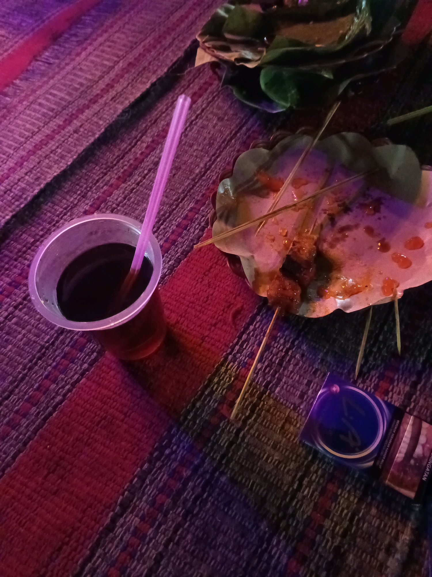 A still life image captures a close-up view of a meal and a drink on a woven mat with alternating bands of dark red, pink, and purple. To the left, a clear plastic cup filled with a dark liquid, likely iced tea, contains a pink straw. In the center right, a used paper plate holds the remains of several skewers of food, some with sauce and visible meat. Two wooden skewers are placed horizontally on the plate, and another two stick out vertically. To the right of the plate is a partially visible cigarette pack, predominantly blue with a circular emblem and text in Indonesian that appears to be a health warning. The lighting in the scene is dim, with a purple and pink cast, suggesting it may be evening or indoors with colored lights. The setting is outdoors on a woven mat, and the presence of food and drink suggests a casual dining or social gathering. The location is identified as Ngasem, Indonesia.