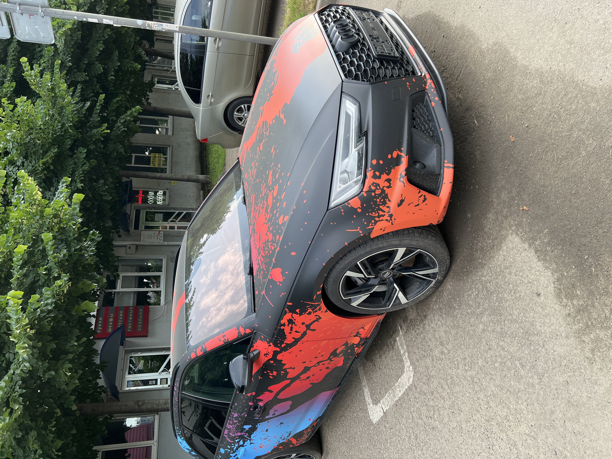 The image captures a black Audi A4 Avant with a distinctive red and blue splatter paint job, parked on a paved street in front of a row of shops. The car is the primary subject, with its custom vinyl wrap drawing significant attention. The shops in the background appear to be commercial establishments, with signage visible in Russian. One sign, in particular, lists "КУХНИ" (kitchens), "ШКАФЫ" (wardrobes), "ГОСТИНЫЕ" (living rooms), and "СПАЛЬНИ" (bedrooms), suggesting furniture or home goods stores. Another smaller sign displays a phone number and the word "АРЕНДА" (rent). The environment suggests an urban setting, likely a commercial district within Krasnodar, Russia, given the Russian language signage. The car is parked in what appears to be a designated parking spot, indicated by white lines on the asphalt. The overall lighting suggests it is daytime, with the sky reflected in the car's windows. Another light-colored sedan is visible parked behind the Audi.