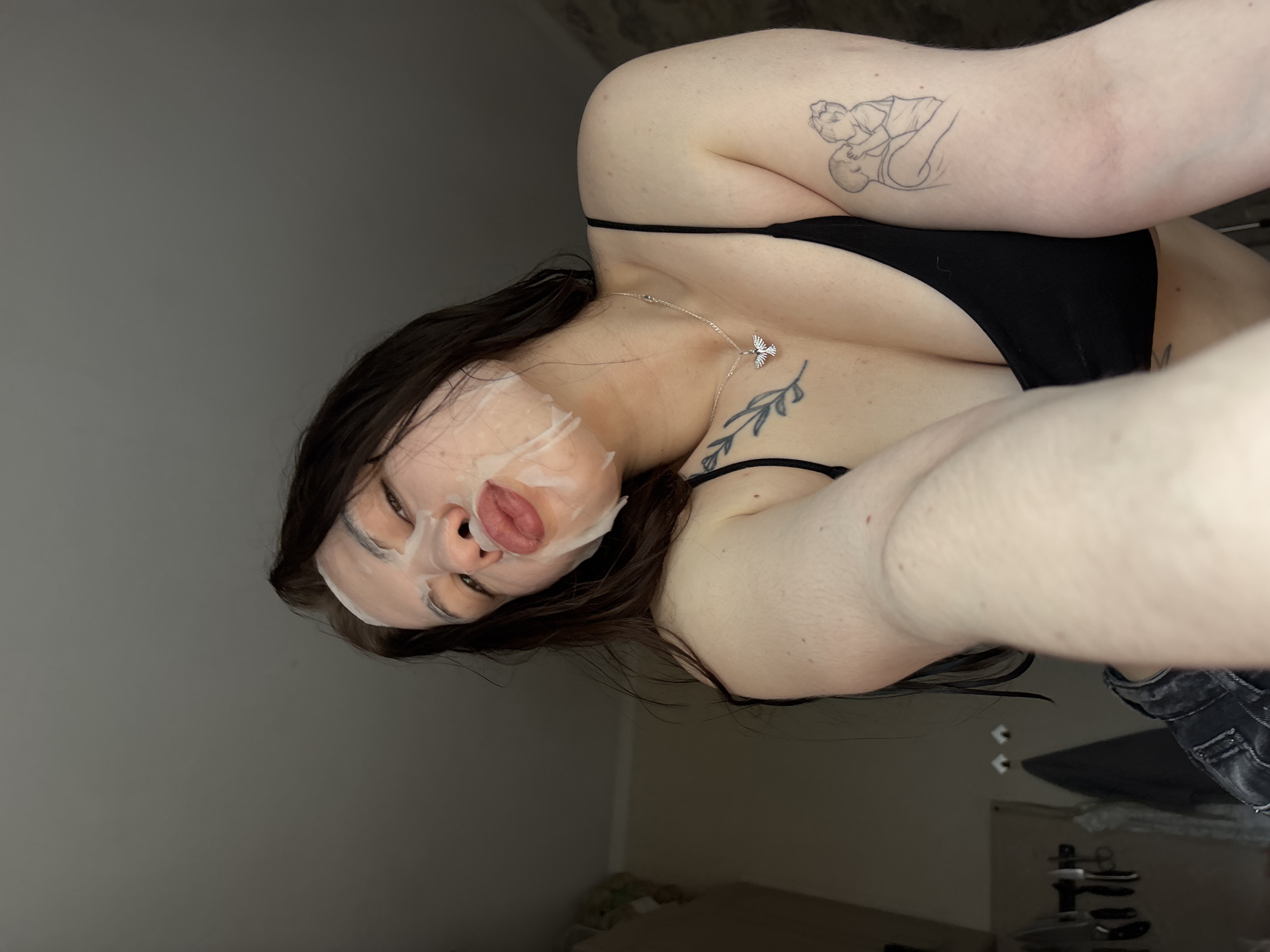 A woman with long brown hair, wearing a black top and a sheet mask on her face, is positioned indoors. She has tattoos on her upper arm and chest. A silver necklace with a bird pendant is visible around her neck. The background features a wall and furniture, suggesting a residential setting. The location in the city of Perm, Russia is not discernible from the image.