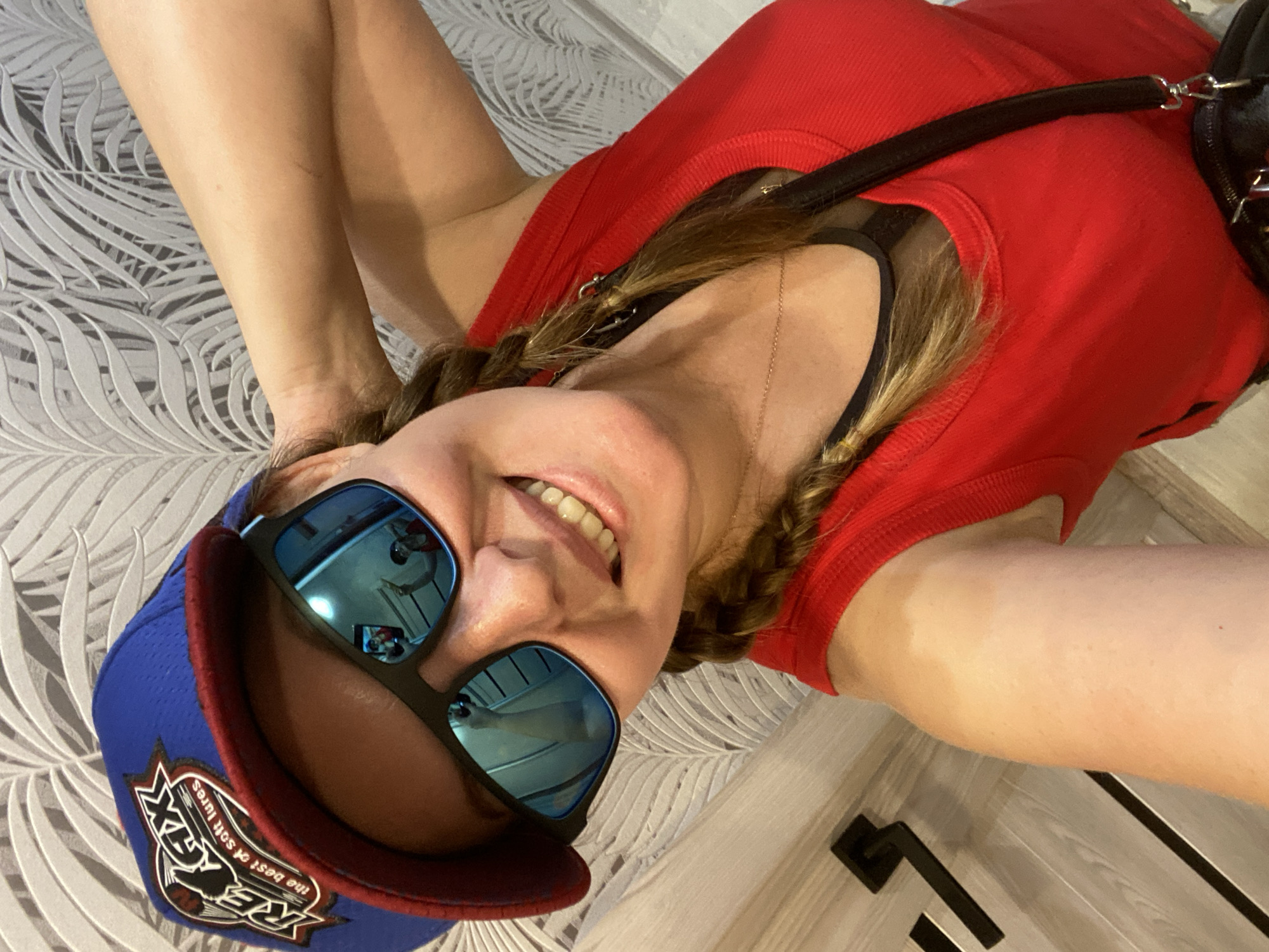 In Noviy, Russia, a woman takes a selfie in a room with a palm leaf patterned wallpaper. She is wearing a blue and red baseball cap with the "RELAX" logo, mirrored sunglasses reflecting an image of herself and a wall, and a red tank top with a black mesh accent underneath. Her right hand is behind her head, and her hair is braided into two pigtails. She is smiling, showing her teeth. A black handbag strap is visible across her shoulder. The background features a door with black handles.