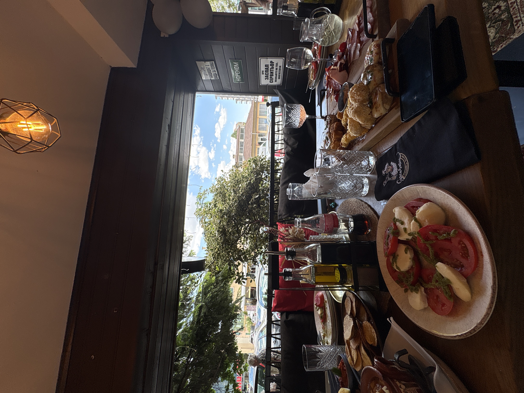 Here is a detailed description of the image:
The image appears to be taken in a restaurant in Sofia, Bulgaria. A window is visible with writing in Cyrillic. A sign with Cyrillic reads, "ДРУГАРИ - БАКШИШЪТ Е ОБИДА ЗА ТЕЛЯКА!" ("Friends - tip is an insult to the tellyak!"). Another sign reads "НОРЕНОМ" ("HORENOM"), followed by "ПРИ ЗАСПАЛ СЕРВИТЬОР ПОЗВЪНЕТЕ НА ТЕЛ: 0887072814" ("If the waiter is asleep call this number").
The focus is on a table laden with food, with an array of dishes including what seems to be a caprese salad (tomatoes and mozzarella), fried chicken, cured meats, and possibly some kind of potato chips. There are also multiple glass bottles, perhaps with dressings or condiments, and several decorative glasses. A tablet is propped up at the edge of the table.
The scene is set in a restaurant or outdoor dining area, with the window looking out onto a street scene with trees, parked cars, and buildings in the background.