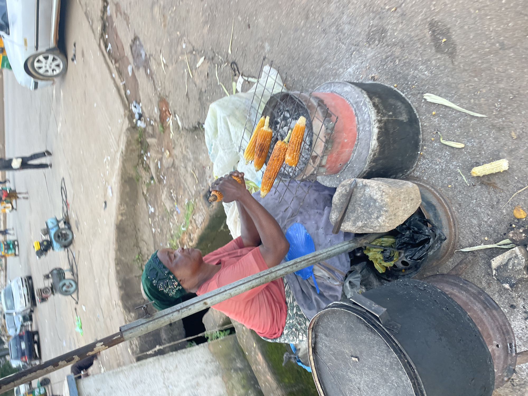 A woman in a green headscarf and an orange t-shirt is roasting corn on a makeshift grill. She is seated on the ground, with a large black pot to her left and a charcoal grill in front of her. The grill has several cobs of corn cooking on it. The scene appears to be a street in Jos, Nigeria, with a paved road and some debris visible. In the background, several vehicles, including a white sedan and a tuk-tuk, are parked or moving. The overall impression is a busy street scene in an urban environment.