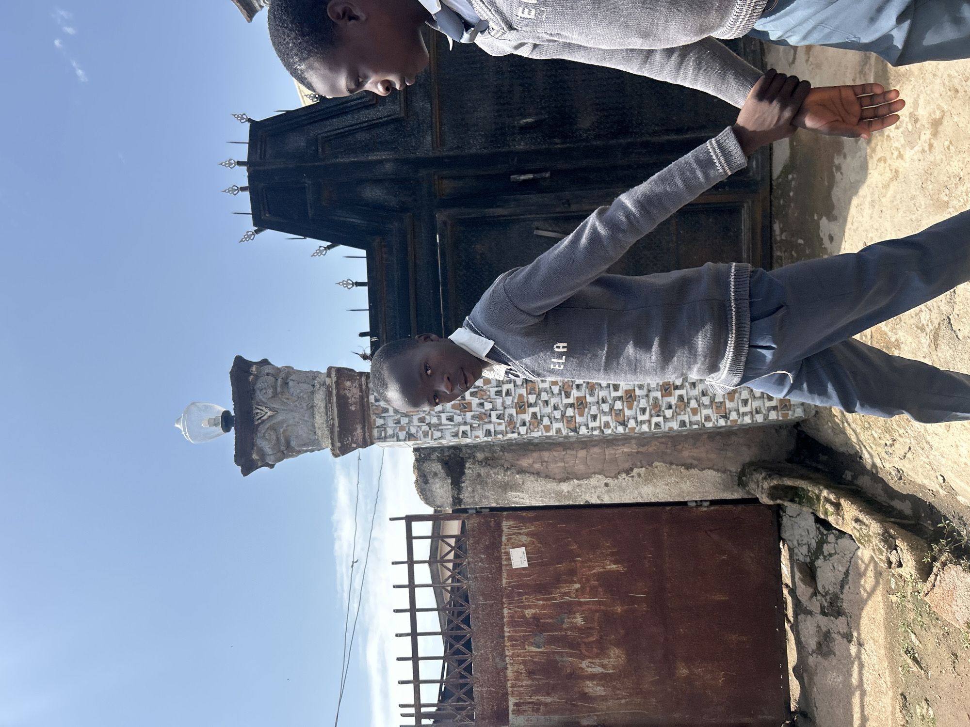 The image depicts two young boys, appearing to be of African descent, standing in an outdoor setting. They are both wearing grey sweaters over white collared shirts and blue trousers, suggesting they might be in school uniform. One boy is in the foreground, looking towards the camera with a neutral expression, while the other is partially visible in the background, reaching out to the first boy's hand. They appear to be in the process of a handshake or a similar gesture.
The background features a dark, ornate metal gate with decorative spikes. To the left of the gate, there is an old stone pillar with carvings and a glass lamp fixture. The wall behind the pillar has a patterned tile design. Further to the right, a large, rusty metal door is visible. The ground is a mix of concrete and dirt.
The sky is clear and blue, with a few wispy clouds, suggesting it is daytime. The lighting indicates a bright day, possibly mid-morning or afternoon, with strong shadows cast on the ground. The scene appears to be in an urban or semi-urban area, possibly a schoolyard or the entrance to a compound in Jos, Nigeria. The overall mood is calm and suggests a simple, everyday interaction between the two boys. Visible text includes the letters "ELA" on the sweater of the boy in the foreground.