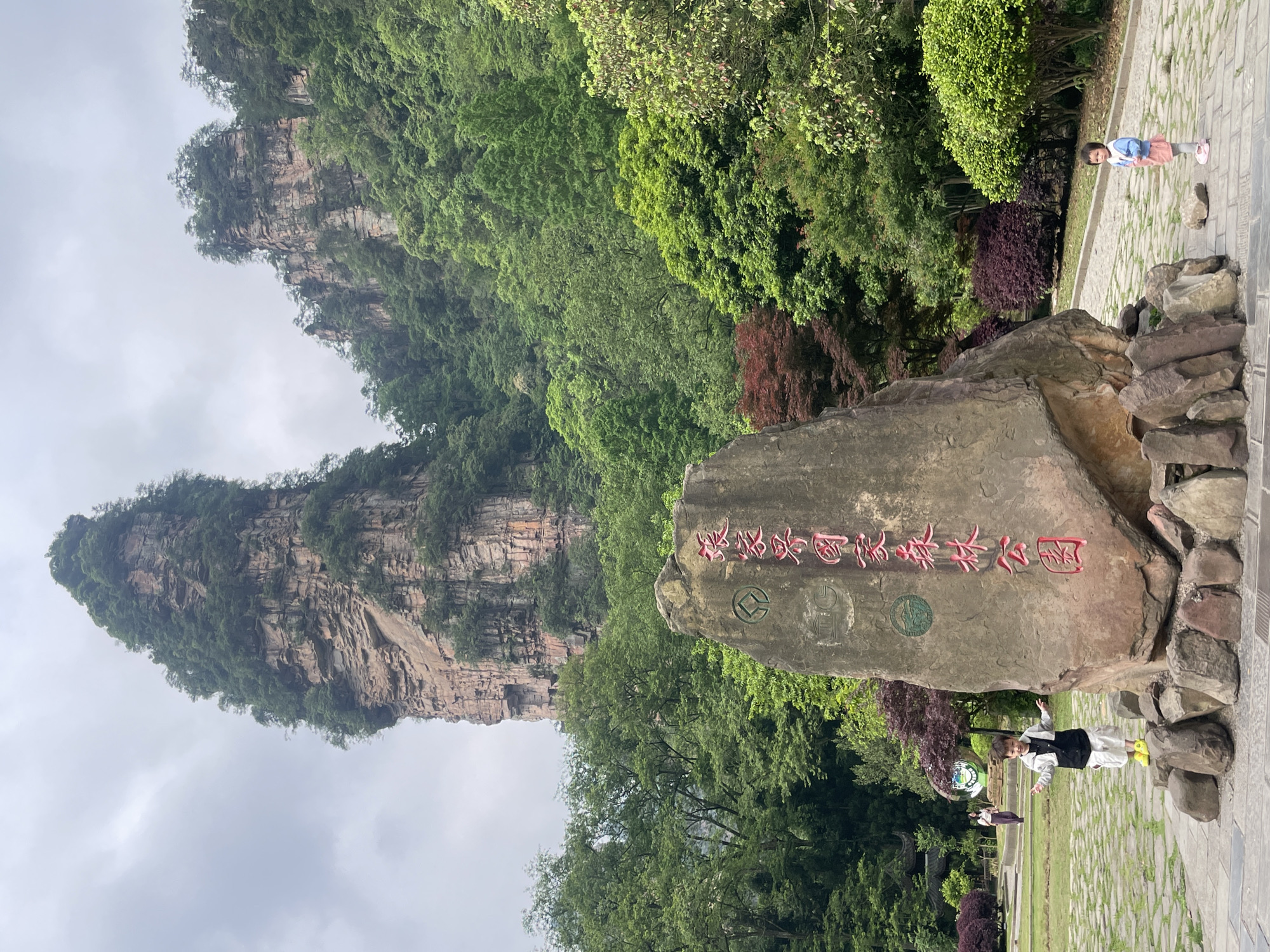 The image shows a scenic view in Luoguta, China. A prominent rock formation rises dramatically in the background, its steep cliffs covered in lush greenery. In the foreground, a large stone boulder serves as a monument, inscribed with Chinese characters in red. Two people are visible in front of the boulder, appearing to be tourists. Further to the right of the boulder, a child stands on the paved path, looking towards the camera. The weather appears overcast, with a cloudy sky above. The scene includes a paved path, various trees, shrubs, and decorative plants.
