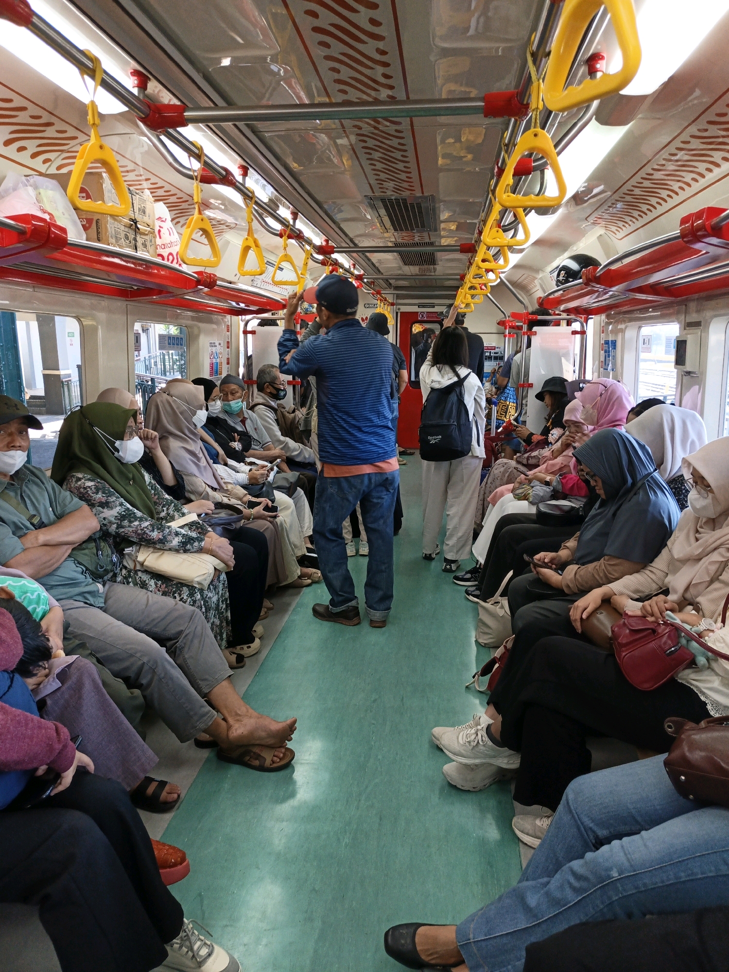 The image depicts the interior of a train car filled with passengers. The passengers are predominantly of Asian descent and are seated on the left and right sides of the aisle. Many are wearing masks, indicating public health precautions. The train car has a light-colored ceiling with red accents and yellow handgrips hanging from yellow poles. The floor is a light green color. The lighting suggests it is daytime, with natural light entering from windows on the sides. Some passengers are looking at their phones, while others appear to be resting or conversing. The overall atmosphere suggests a typical commuter train journey. The mention of Klaten, Indonesia, suggests this is a public transportation scene within that city.